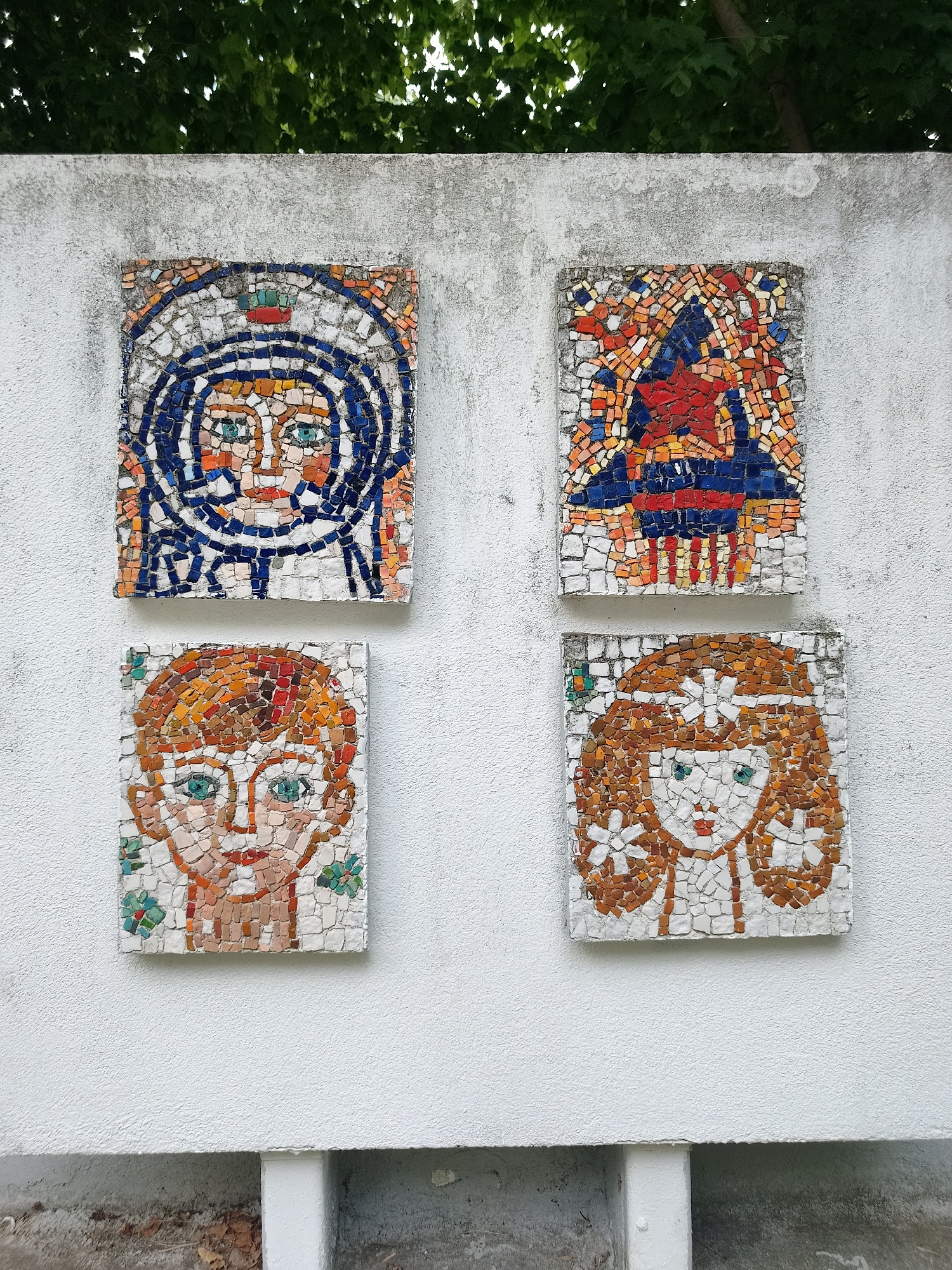 This image displays four mosaic artworks affixed to a white stucco wall. The mosaics are arranged in a two-by-two grid. The top-left mosaic depicts a stylized face, possibly of an astronaut, wearing a helmet with blue and white concentric circles around the face. The top-right mosaic features an abstract design with red, orange, and blue tiles, forming a central red star-like shape. The bottom-left mosaic presents a portrait of a child with orange hair and green eyes, surrounded by small green floral accents. The bottom-right mosaic shows a portrait of a girl with long, wavy brown hair, green eyes, and a floral headdress with white flower-like designs. The wall is textured, and the mosaics appear to be made of small, irregularly shaped tiles. Above the wall, green foliage from trees is visible. The bottom of the image shows a concrete base with some debris and foliage. These mosaics are located on the wall of the former Pioneer Home (Dom na pionerite) in the city of Sandanski, Bulgaria.