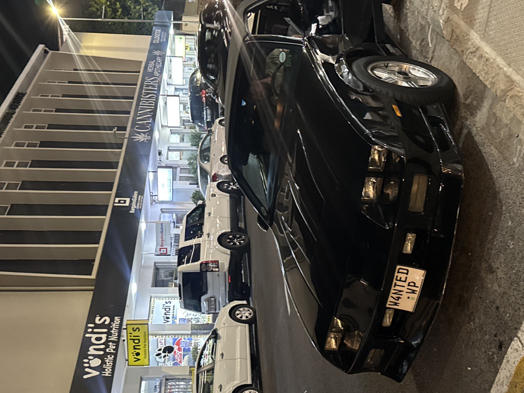 The image is an eye-level, outdoor photograph of a black car parked on a street in Cape Town, South Africa. The car's license plate reads "W4NTED" : WP", and it is parked next to a building with various businesses. The building has signs for "vondi's Holistic Pet Nutrition," "Bargains Galore," and "CANNIBISTERS Herbal Apothecary." There are several cars parked behind the black car. The image was taken at night, and the lighting is provided by streetlights.