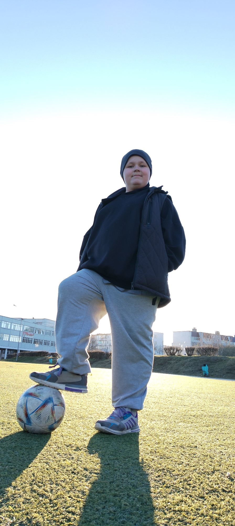 A young person stands outdoors with one foot resting on a soccer ball, on the city of Chaikovsky, Russia. The individual wears a dark-colored beanie, a dark hooded sweatshirt, a black vest, light gray sweatpants, and gray athletic shoes with purple accents. The soccer ball is white with multicolored accents. They stand on artificial turf. Behind them is a building. The sky is clear.