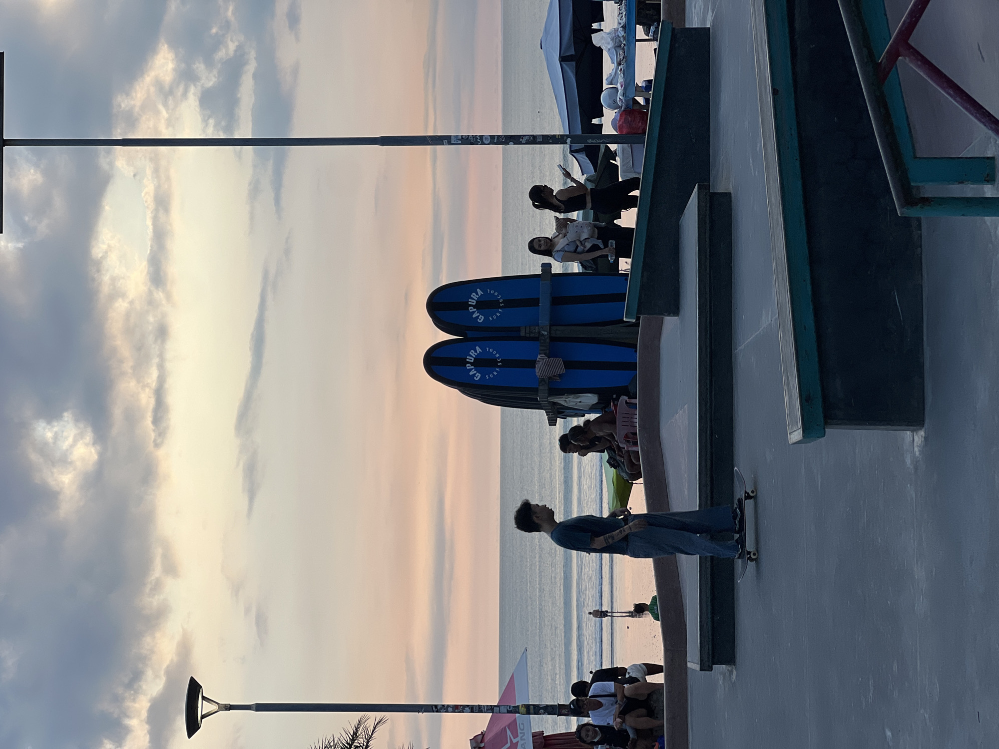 The image depicts an evening scene at a beachside location in Kuta, Indonesia. The sky exhibits a gradient of colors from pale yellow to light pink, suggesting sunset or sunrise. Clouds are visible in the sky.
On the right side of the image is a section of a concrete skate park. A man in jeans and a dark shirt stands on a skateboard. To his left, several people are sitting or standing, observing the beach.
Further to the left are two blue surfboards mounted on a rack. Beyond the surfboards, people are sitting or standing along the beach.
A light post extends from the bottom left to the upper left, running vertically. Another light post is visible in the lower left corner.
The beach is visible in the background.