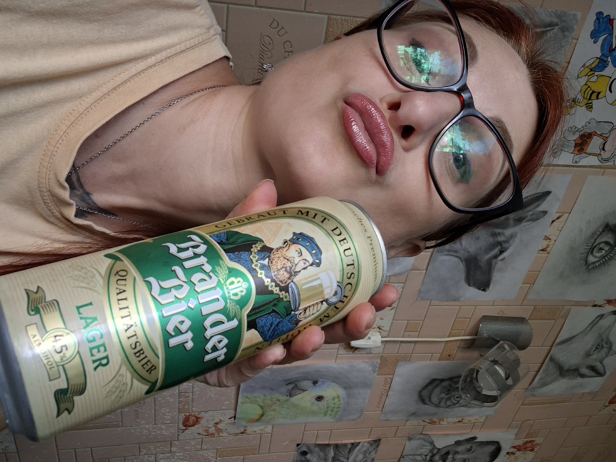 A woman with auburn hair and glasses holds a can of "Brander Bier" while looking at the camera. She is wearing a light-colored t-shirt and a silver necklace. Behind her, the wall is decorated with various drawings, including portraits of animals and a baby. The scene appears to be indoors, possibly in a room in the city of Myadzyel, Belarus.