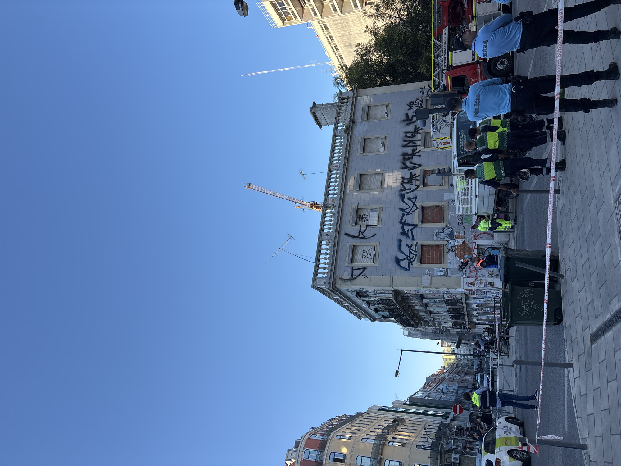 The scene is in Lisbon, Portugal. A street is cordoned off with police tape, with a line of Portuguese police officers and security personnel standing behind it. Behind them is a fire engine and a building covered in graffiti. In the distance, buildings line the street. A construction crane is visible behind the graffitied building. The sky is clear and blue. A police car is parked nearby. Two large, dark green trash cans are positioned at the curb.