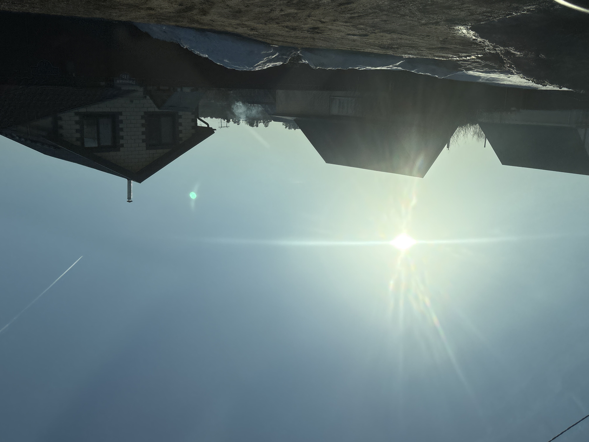 The image, rotated 180 degrees, shows a view of Chaikovsky, Russia under a bright, sunny sky. The sun dominates the upper portion of the image, with strong light flares. Below, there are several houses visible, their roofs angled. A white contrail of a high-flying jet cuts across the lower left portion of the sky. Some snow is on the ground, and what appears to be smoke is rising from one of the house's chimneys. The foreground likely consists of more ground.