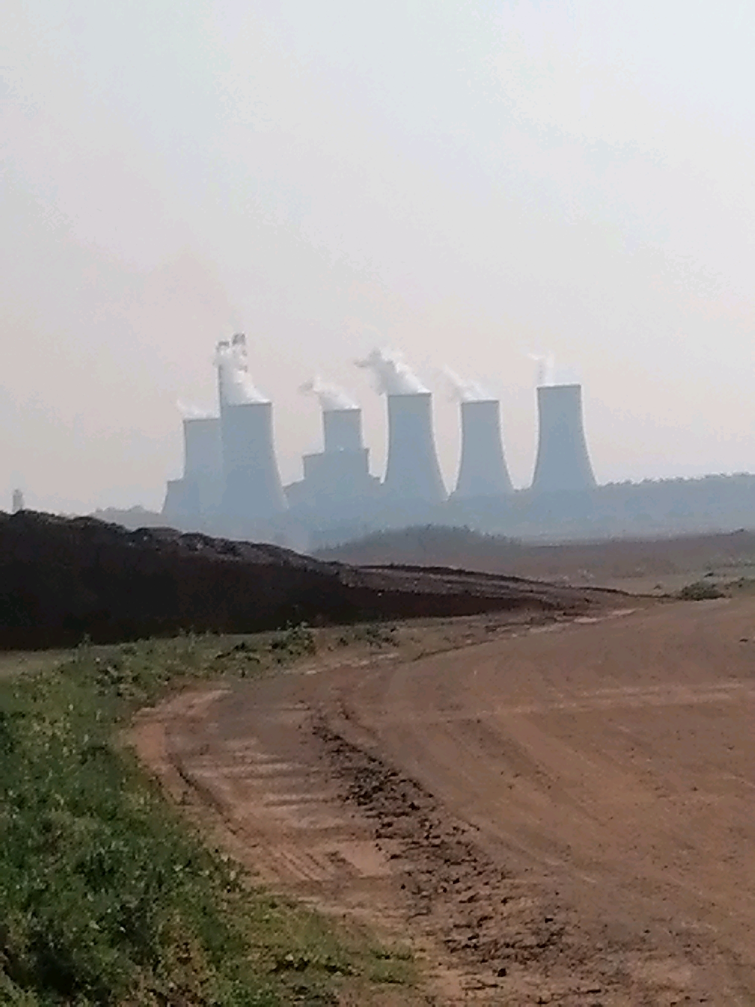 This image depicts a wide view of the coal-fired power stations within the Midvaal Local Municipality, South Africa. In the foreground, a dirt road curves from the bottom left to the middle right of the frame. To the left of the road, a patch of green grass and sparse vegetation is visible. Behind the road and vegetation, a large pile of dark brown material, likely coal or slag, dominates the middle ground. In the background, several large, white cooling towers of a power plant rise above a hazy horizon. White plumes of steam or smoke emanate from the tops of these towers, indicating ongoing industrial activity. The sky is overcast and hazy, creating a muted color palette for the scene. The overall impression is one of industrial landscape under a hazy sky.