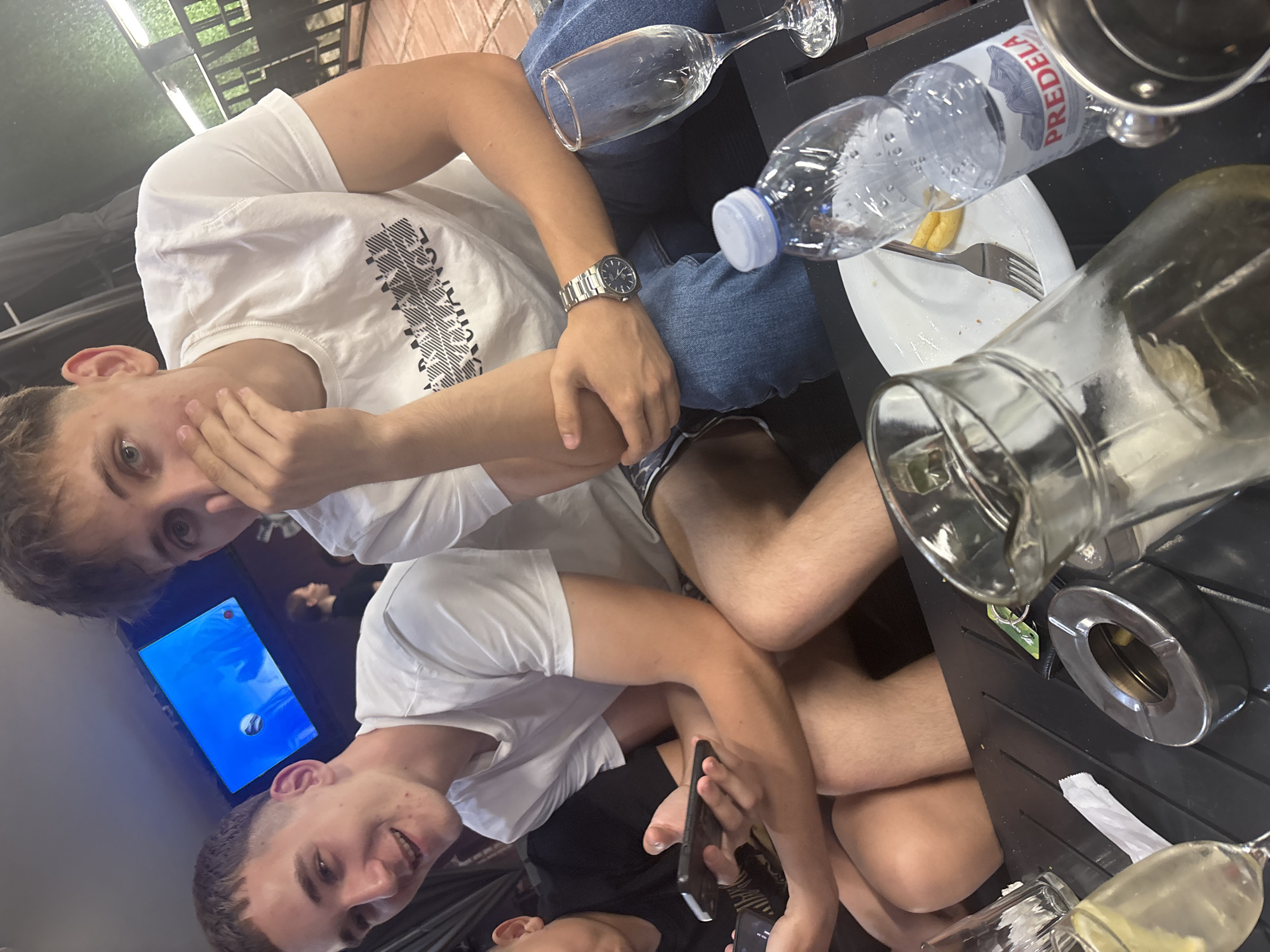 A casual gathering is depicted at an outdoor or semi-outdoor cafe or restaurant, likely situated in Blagoevgrad, Bulgaria, identifiable by the "PREDELA" brand mineral water bottle. The foreground features a dark-colored table holding a clear plastic bottle of Predela mineral water with a blue cap, a glass pitcher containing a light-colored liquid, two empty stemmed glasses, a metal ashtray with remnants, a white plate with a fork and yellow food residue, a green keychain, and a white paper napkin.
Three young males are visible. The male on the left, with short dark hair and a white t-shirt, smiles directly at the camera while holding a dark smartphone. Adjacent to him, a male with light brown hair, blue eyes, and a white t-shirt bearing "ARMANI EXCHANGE" text covers his mouth with his left hand, looking towards the camera. He wears a silver-toned wristwatch on his left wrist and blue jeans. A third male's arm and black t-shirt are partially visible on the far left, also holding a smartphone. In the background, a female with dark hair and a dark top is seen to the right of the center male, positioned beneath a wall-mounted television displaying a blue screen with a white spherical object. The environment includes dark fencing, green foliage, and dark fabric coverings.