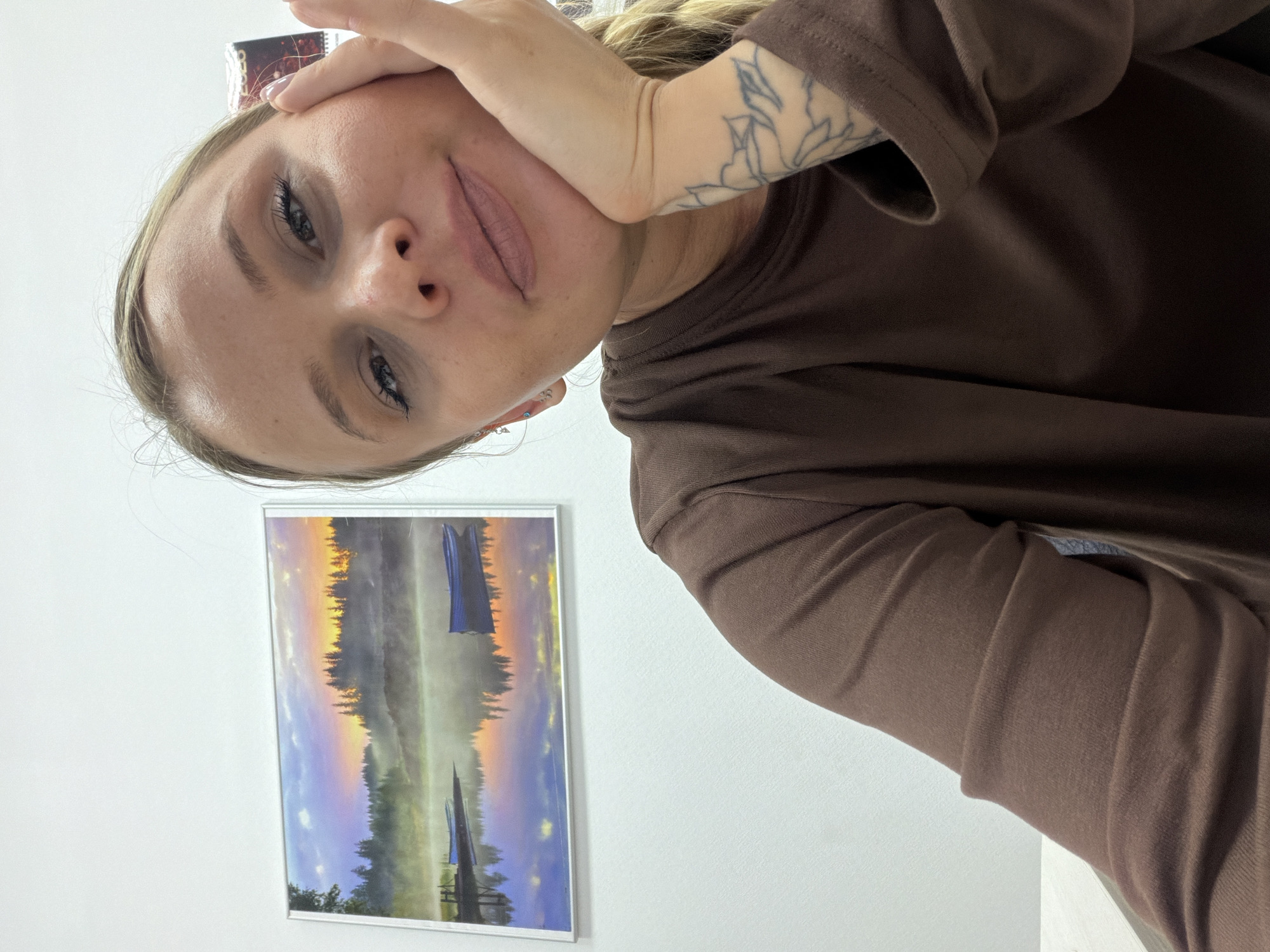 A woman with blonde hair and a brown long-sleeved shirt poses indoors. She rests her head in her hand, revealing a tattoo on her forearm. Behind her is a framed picture of a lake landscape at sunset or sunrise. The walls are white. There is no verifiable information to confirm the location is Sarapul, Russia.