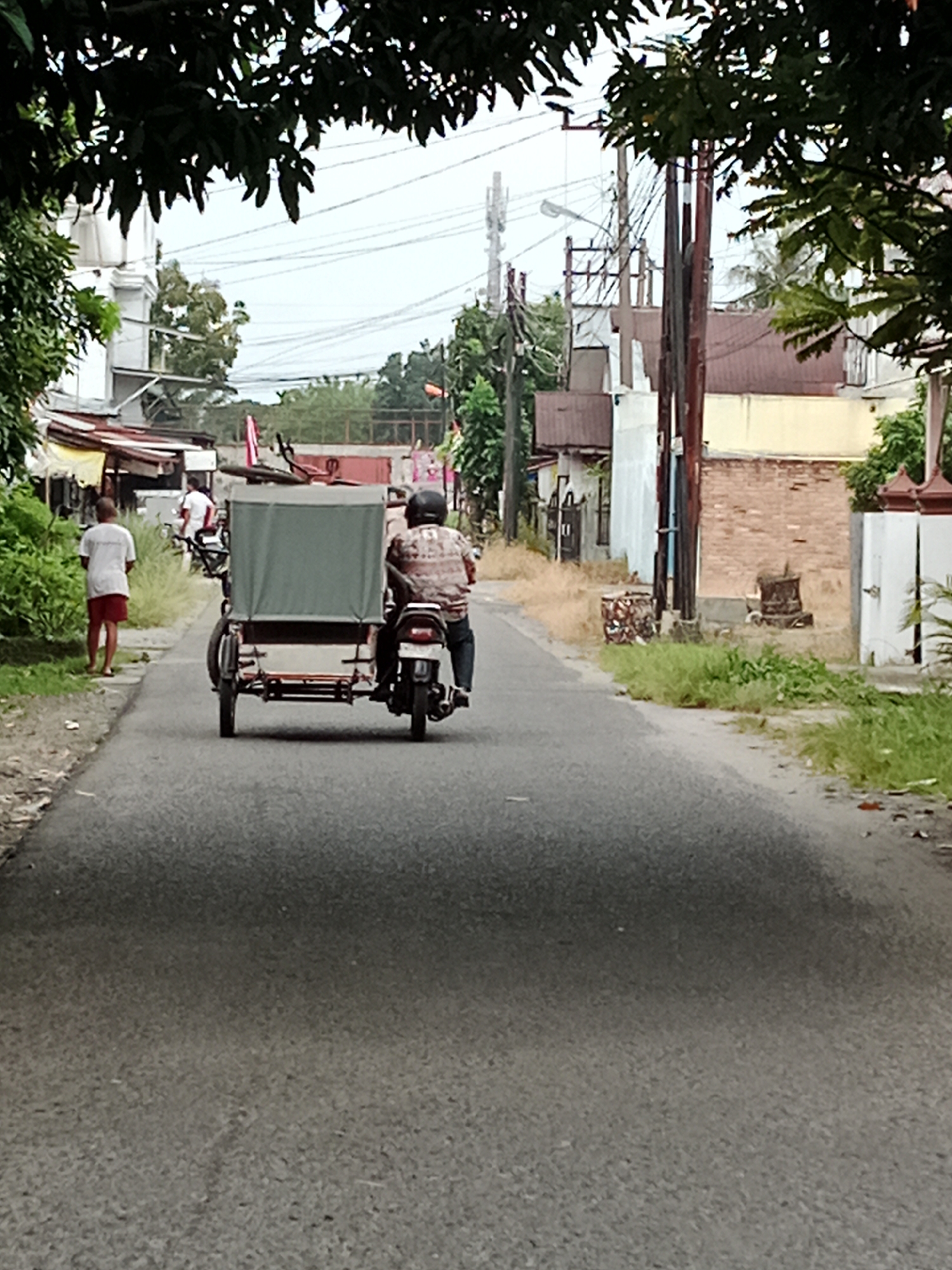 In the city of Binjai, Indonesia, a tricycle driver is seen operating a three-wheeled motorcycle with a passenger cabin on a paved road. The tricycle is gray and has a fabric covering over its passenger area. The driver, wearing a checkered shirt and dark pants, is seated on the motorcycle, while the passenger's side of the tricycle is obscured from view. A young boy in a white shirt and red shorts stands on the side of the road. The scene depicts a typical street in Binjai, with buildings and vegetation visible in the background, as well as utility poles and electrical wires crisscrossing overhead. The road surface appears worn, and the surrounding environment suggests a warm, tropical climate.