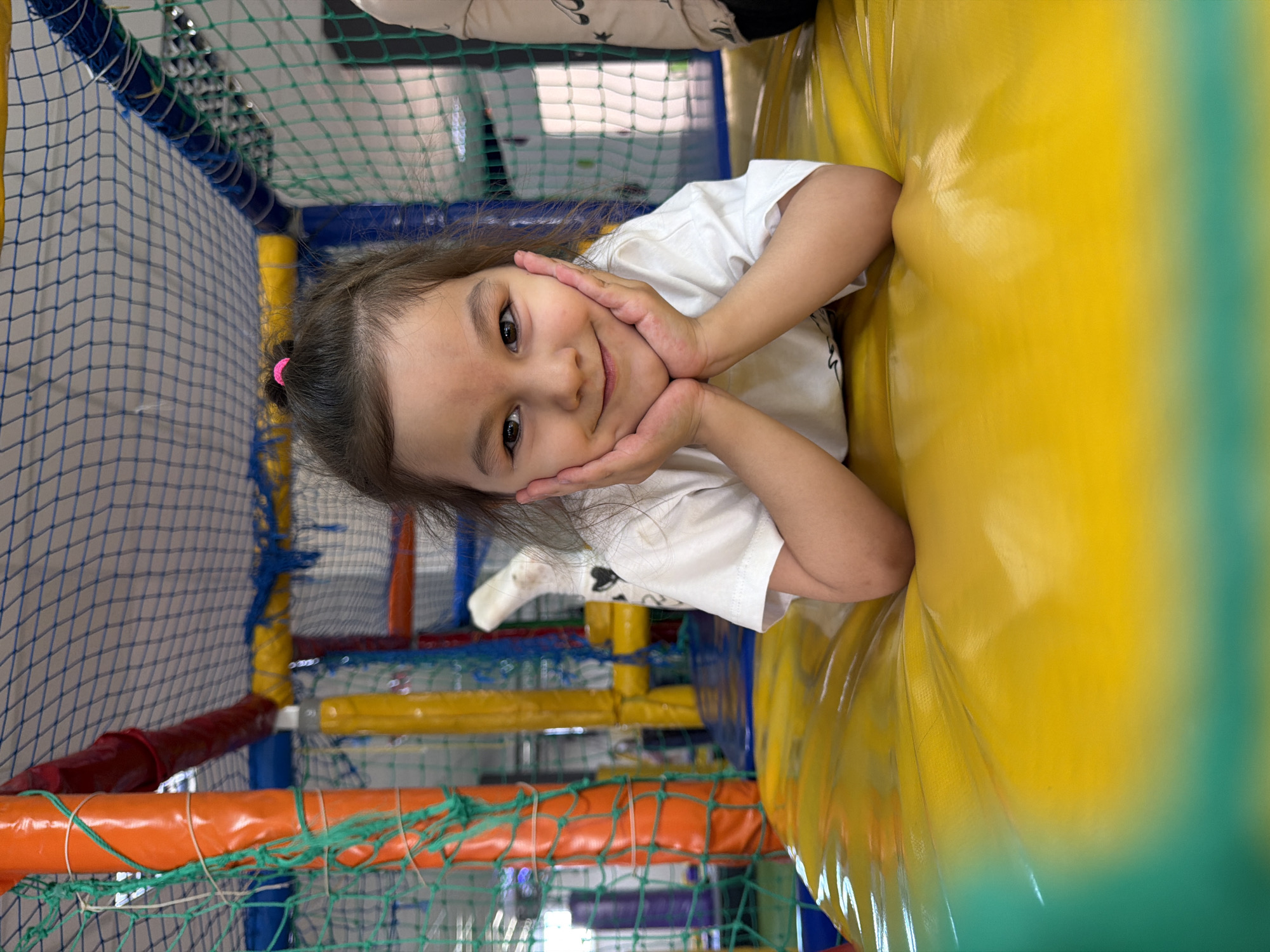 A young girl with brown hair tied in a small pink ponytail is smiling, with her hands resting under her chin. She is positioned within an indoor playground, leaning against a yellow padded structure. The playground features multicolored barriers made of netting, tubes and padding in red, orange, yellow, and blue. The girl is wearing a white T-shirt with a small print design. The background appears to be a play area enclosed by netting. This scene is located in Chaikovsky, Russia.