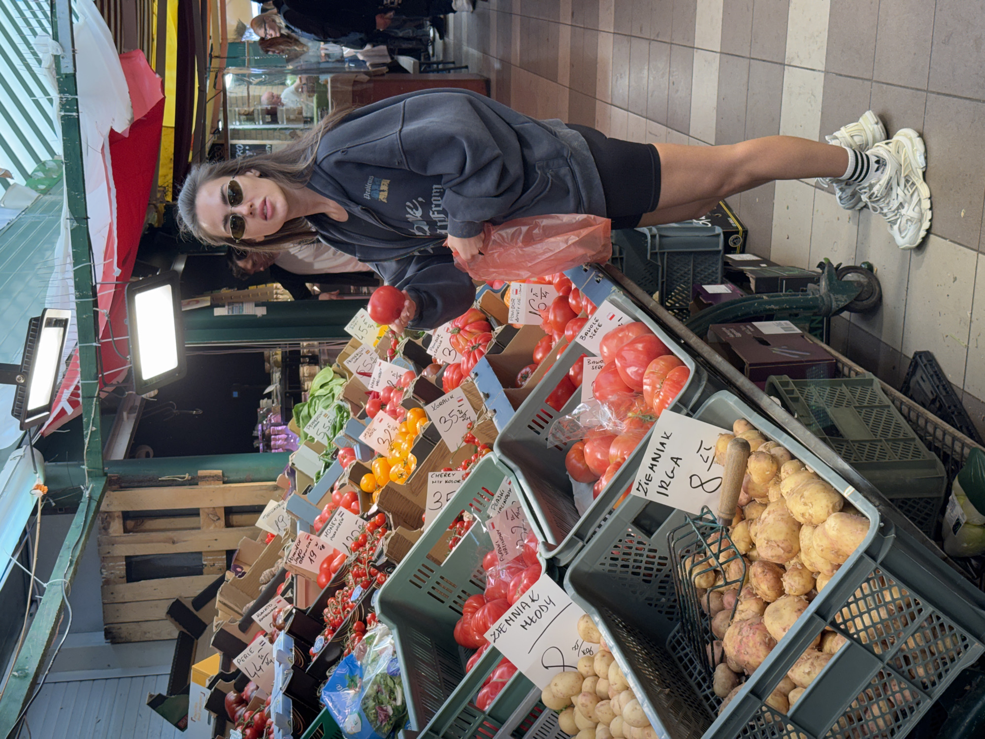 The image depicts a woman in sunglasses and athletic attire selecting a tomato from a produce stand at a marketplace in an urban environment. She is surrounded by crates filled with various vegetables, including tomatoes of different varieties and potatoes, each labeled with price tags written in Polish. The overhead structure of the market is visible, composed of wooden beams, awnings, and lighting fixtures. Shoppers are vaguely visible in the background, adding to the bustling atmosphere of the market.
