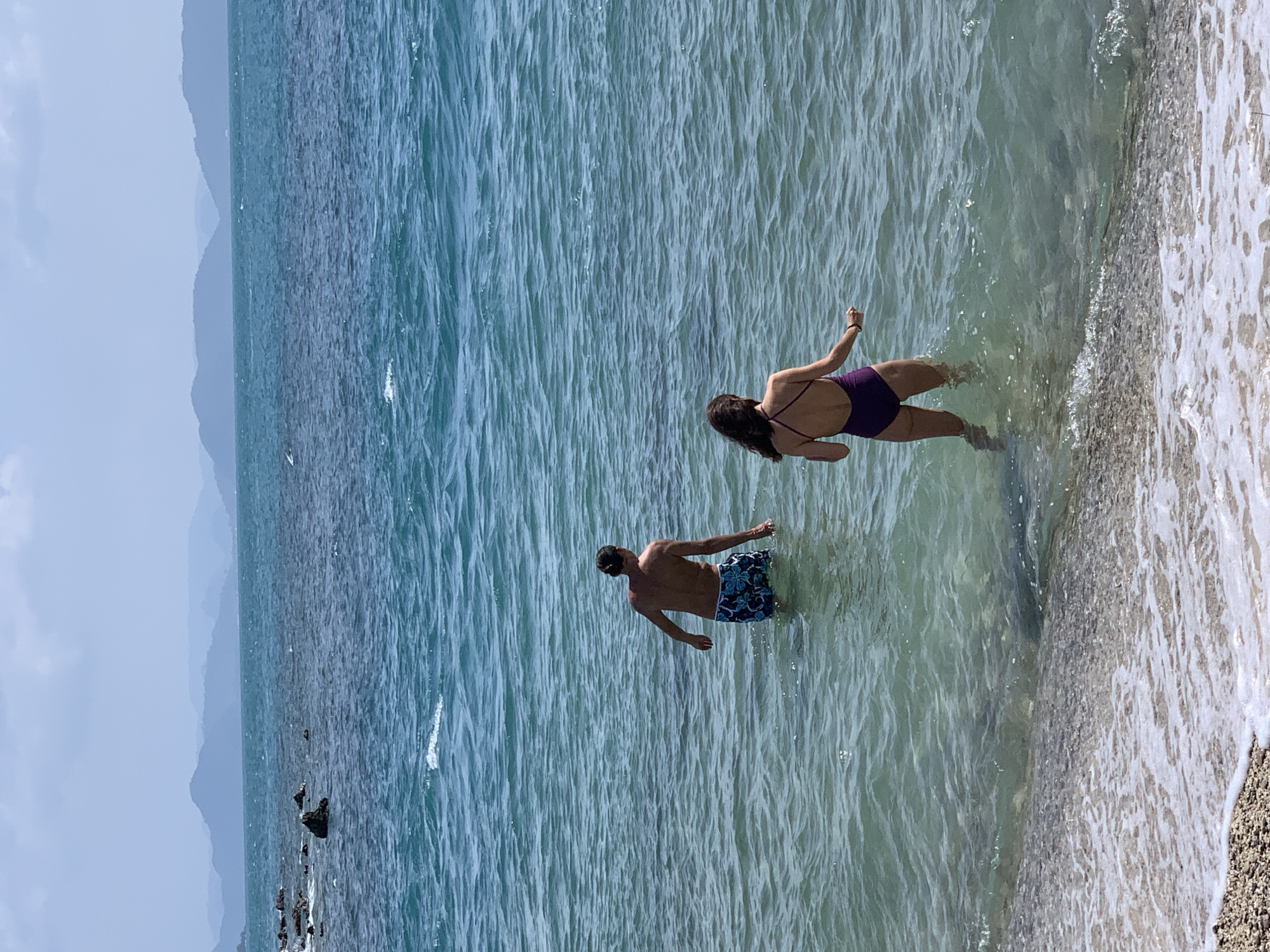In the city of Unknown, Thailand, a man and a woman stand in the shallow turquoise waters of the ocean. The man, facing away from the camera, wears patterned swim trunks and is partially submerged. The woman, also with her back to the camera, wears a dark purple one-piece swimsuit and is wading in the water. The water is clear, with visible ripples and waves. In the background, layers of hazy blue mountains rise against a pale blue sky. The foreground shows a pebble beach with white foam from the breaking waves.