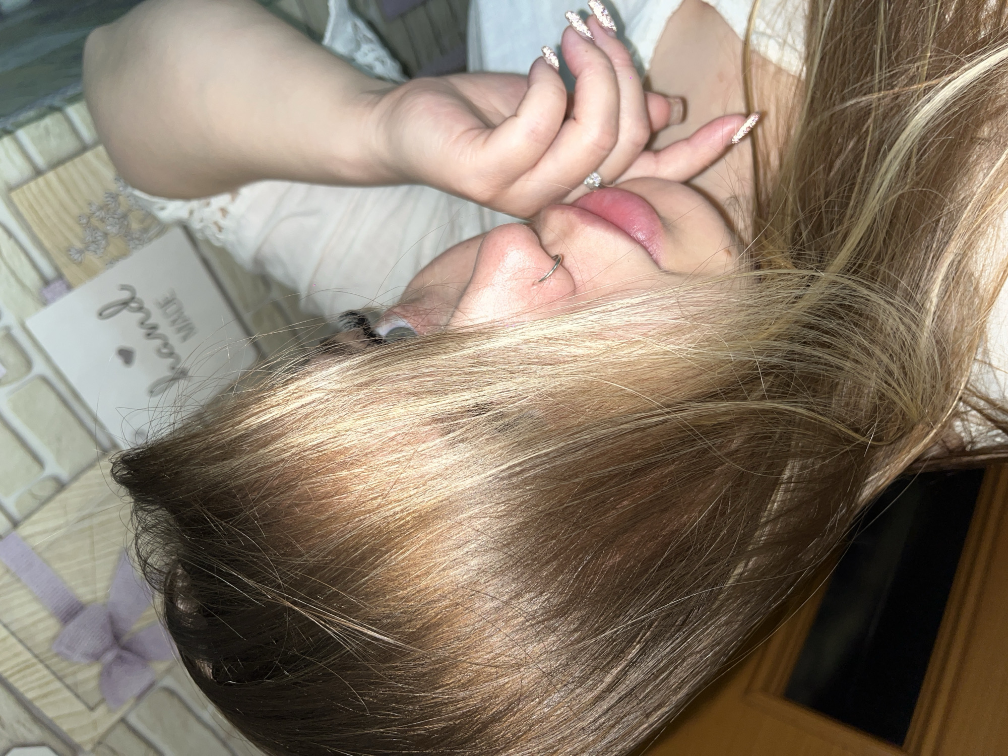 A close-up, angled shot depicts a person with long, blonde hair with dark roots, lying down, likely on a bed. The person has a nose ring and is wearing white clothing. Their hand with a diamond ring and glittery nails is near their face, touching their lips. A "hand MADE" card with a light purple ribbon on a tiled surface is partially visible in the corner. The environment is dimly lit with a wooden door in the bottom corner. The location in the city of Port-Katon, Russia cannot be determined.