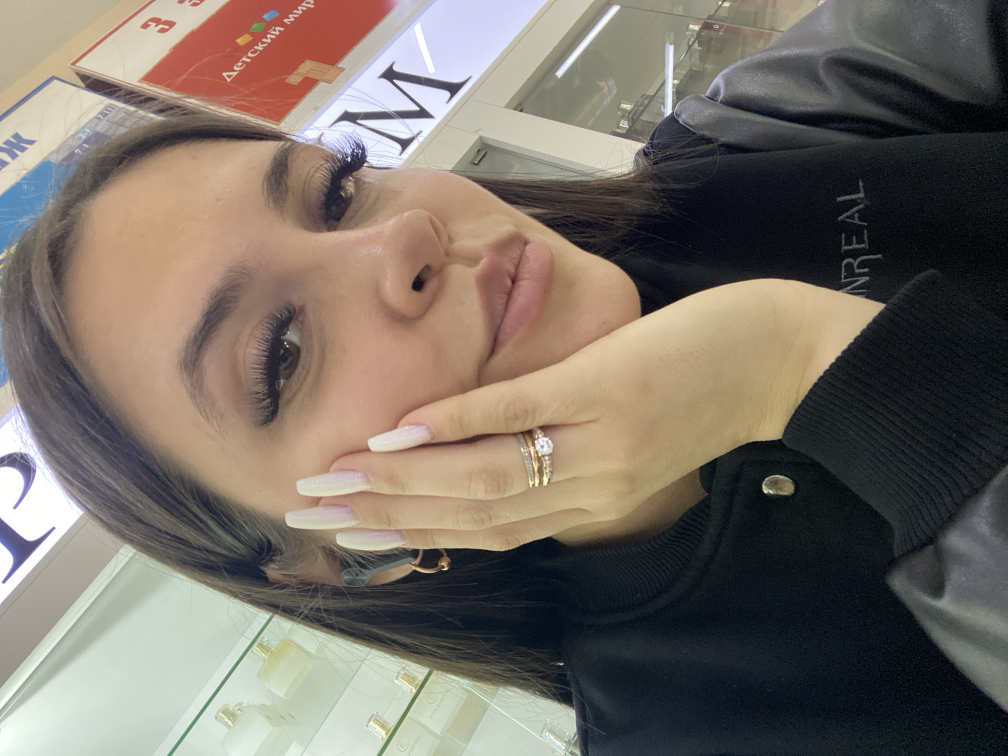 A young woman is featured in a close-up selfie, taken at an angle, with her left hand posed gracefully against her chin. She looks directly at the camera with a soft, neutral expression. Her long, dark hair frames her face, and she has well-defined eyebrows and striking, long eyelashes. Her lips are a natural pink hue, and her skin is fair.
She is dressed in a black long-sleeved top or jacket, which features a ribbed collar and cuffs, and potentially contrasting material on the sleeves. The word "UNREAL" is embroidered in white on her garment. Adorning her left ring finger are three rings: a central gold-toned ring with a prominent clear stone and a paved band, flanked by two thinner gold-toned bands. She wears a small gold hoop earring in her right ear, and her long nails are manicured with a shimmery white-to-pink ombré polish.
The setting appears to be an indoor commercial space, likely a shopping mall or store, characterized by bright artificial lighting and display cases with various bottles, possibly perfumes. Prominent signage in the background includes a red sign with white Russian text that reads "Детский мир" (Children's World), and a large white sign with a dark, stylized letter "M". The presence of "Детский мир," a well-known Russian chain, confirms the location as Russia, consistent with the provided context of Chaikovsky.