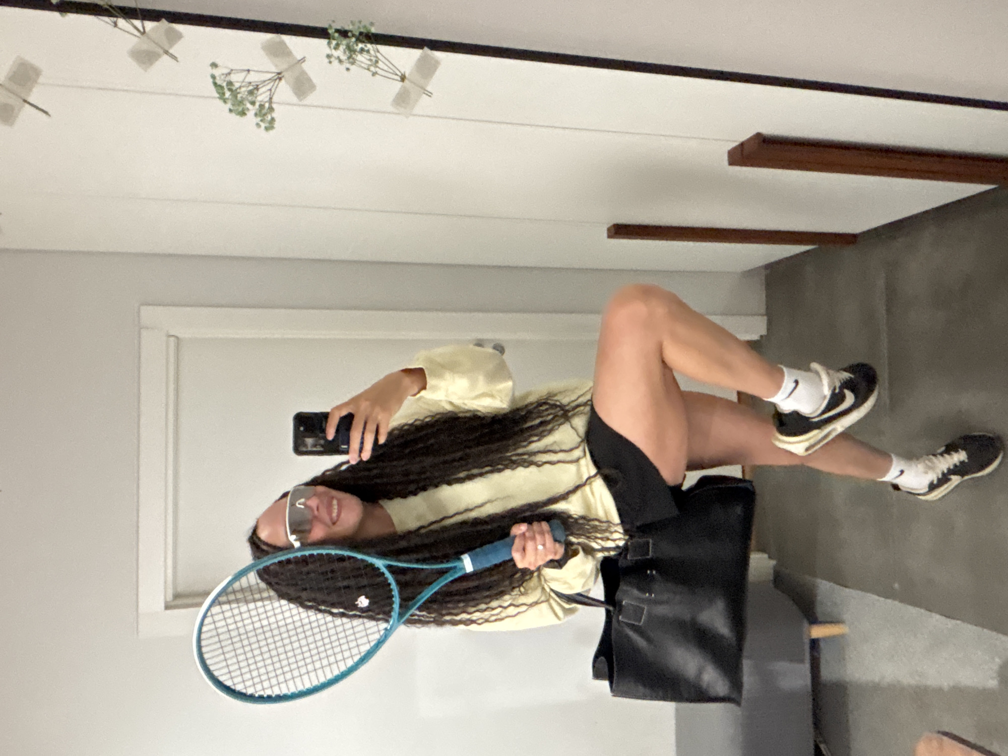 The image, captured in Chaikovsky, Russia, features a young woman taking a mirror selfie indoors. She is smiling broadly, wearing large white-framed athletic sunglasses. Her long, dark, wavy hair falls past her shoulders over a light yellow long-sleeved top and black athletic shorts. On her feet, she wears white socks and black athletic shoes with white soles, identifiable by the visible "Nike" logo.
In her left hand, she holds a black smartphone, seemingly capturing the reflection. Her right hand grips a vibrant blue tennis racket with white strings and a blue grip. A black tote bag rests on the grey floor beside her.
The background shows a minimalist room with white walls and a grey floor. Decorative elements include several small sprigs of green foliage, possibly baby's breath, neatly taped to the upper wall or ceiling. Dark wooden beams or planks are also visible on the wall, contributing to a clean and modern aesthetic. The scene suggests a casual moment, perhaps before or after a tennis activity, or simply a stylish pose within a contemporary interior.