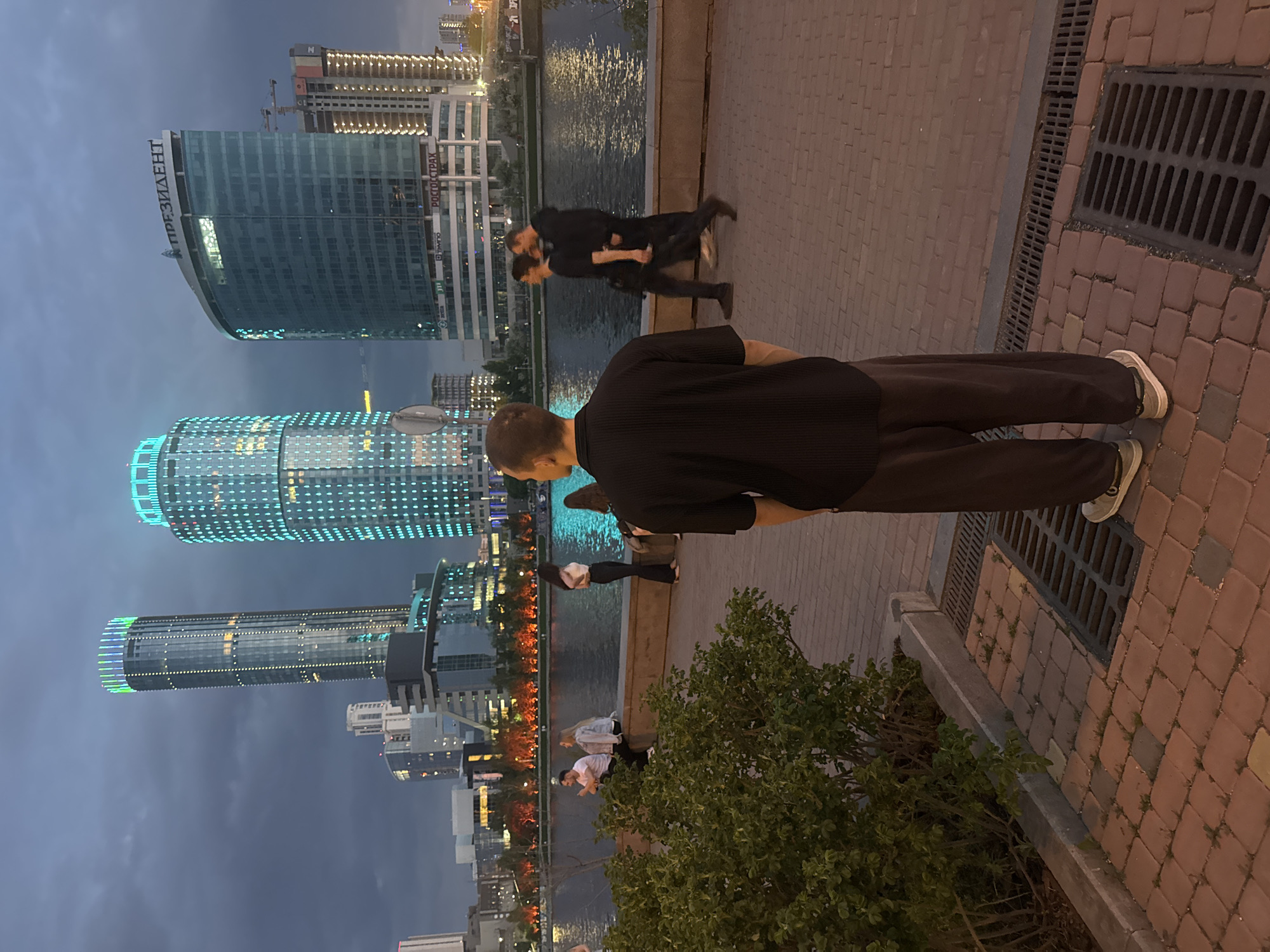 The image depicts a nighttime scene in Yekaterinburg, Russia, along the Iset River embankment. In the foreground, a person in a black shirt and brown pants stands near a bush. Beyond the person, two men are physically interacting, possibly fighting or play-fighting, along the embankment. Several other individuals are visible along the riverbank, some standing and others sitting. The river reflects the lights from the city buildings. Prominent buildings in the background include the "President" office building and other high-rise structures with decorative lighting. The pavement is made of red bricks, and there's a drainage grate near the edge of the sidewalk.