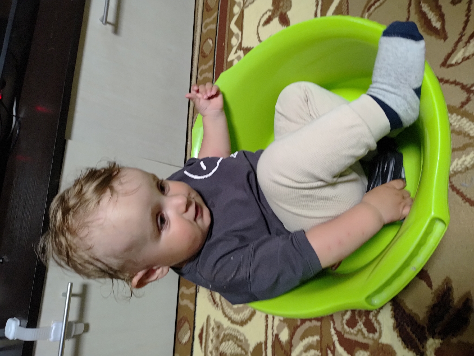 A young child is seated in a green plastic tub on a rug. The child wears beige pants, a grey long-sleeved shirt with a white smiley face, and a grey sock with a dark blue toe. The rug is patterned in brown, beige, and white. A light-colored cabinet with metal handles is partially visible to the left. The scene is located in Марковский, Russia.