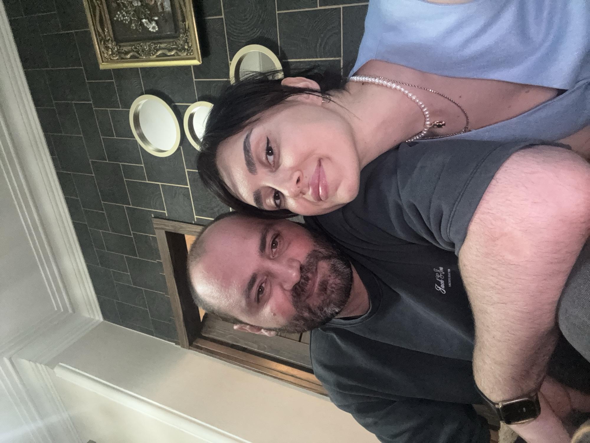 Two people are captured in a close-up, indoor photograph. The location appears to be a room with dark gray tiled walls, adorned with a framed picture and decorative mirrors. The woman is fair-skinned with dark hair, wearing a light blue top and a pearl necklace. She is positioned leaning onto the man. The man has a bald head with a beard. He is wearing a dark gray shirt. A watch is visible on his wrist. The overall setting suggests a personal or social context. It is not possible to determine if the location is in Tbilisi, Georgia based on the image alone.