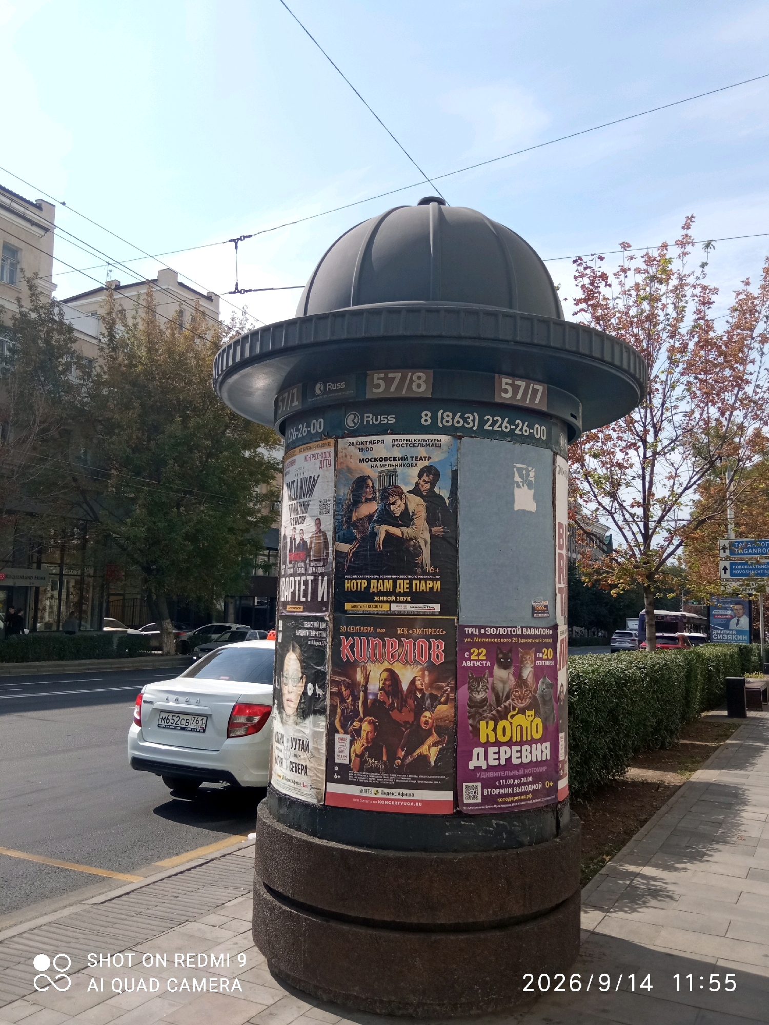 An eye-level, full outdoor shot captures a cylindrical public advertising pillar plastered with various posters. The pillar is situated on a sidewalk next to a street. In the foreground, a white Lada sedan is parked and partially visible. The street features yellow lane markings, and a well-maintained green hedge lines the sidewalk. In the background, buildings and trees contribute to the urban landscape. The sky is clear and blue. The text on the pillar is primarily in Russian, and a timestamp indicates "2026/9/14 11:55". There are no people clearly visible in the shot. This scene is likely in Rostov-on-Don, Russia, based on visual cues common to the city, such as the architectural style of the buildings, typical street infrastructure, and the Russian language used in the advertisements.