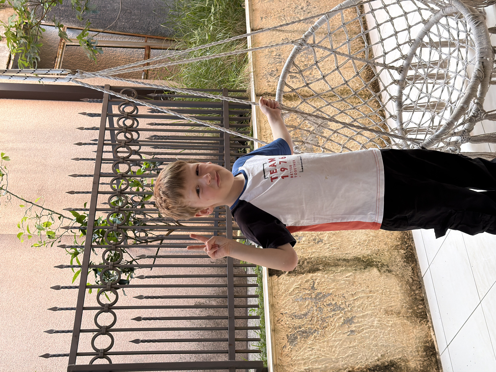 A young boy with light brown hair is depicted standing in an outdoor setting, likely a garden or patio, under bright daylight in Petrovac na Moru, Montenegro. He is facing the camera with a slight smile and making a peace sign with his left hand. His right hand is holding onto the side of a white, netted hanging hammock chair, which occupies a significant portion of the right side of the frame.
The boy is dressed in a white t-shirt with black short sleeves and a red stripe along the bottom edge, paired with black trousers. Visible text on his shirt reads "TEAM 1976 BETTER TOGETHER". Behind him is a dark, ornate metal fence with circular motifs and pointed spikes. Beyond the fence, a textured, light brown or pinkish building wall is visible, along with green foliage and a distant, partially obscured window with a dark frame. The ground beneath him transitions from a pale tiled surface on the right to a rougher, textured ground with some discoloration on the left. The scene suggests a pleasant, sunny day.