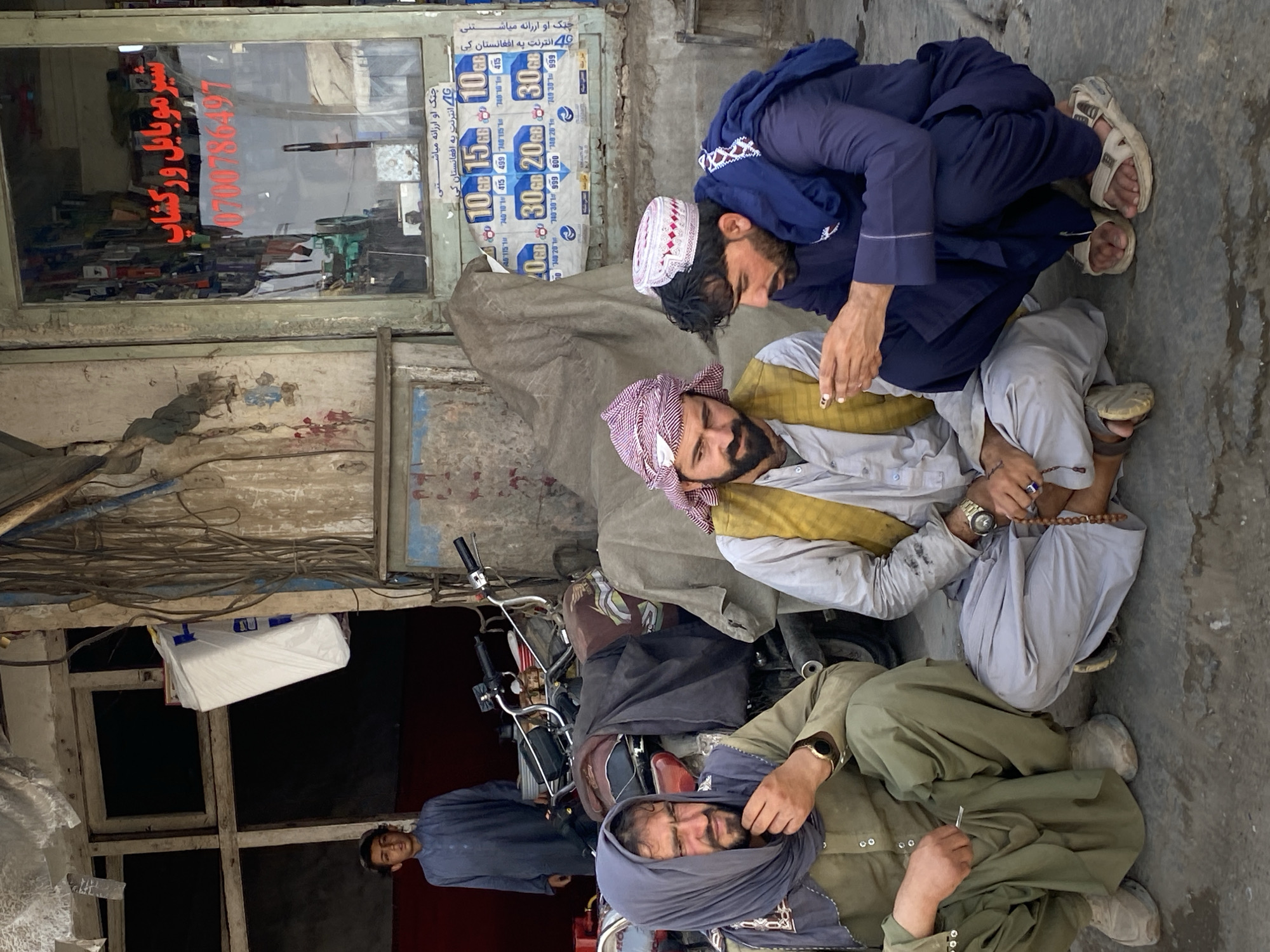 A medium shot captures three men and a boy in an outdoor urban environment in Kandahar, Afghanistan. The men are crouched on a dirt and concrete surface in the foreground, facing the camera. The man on the left wears a green tunic and headscarf, his hand resting on his chin. The central man is dressed in a light-colored tunic, a yellow vest, and a red and white checkered headscarf; he holds prayer beads. The man on the right wears a dark blue tunic and a white cap with red trim, and he is looking down at his hands. Behind the men, a boy in a blue tunic stands in the doorway of a building. A motorcycle is parked behind the central and right-hand men. The background shows a weathered building with signs and wires, suggesting a commercial area. The overall scene depicts a moment of rest or conversation among the individuals in a typical street setting.