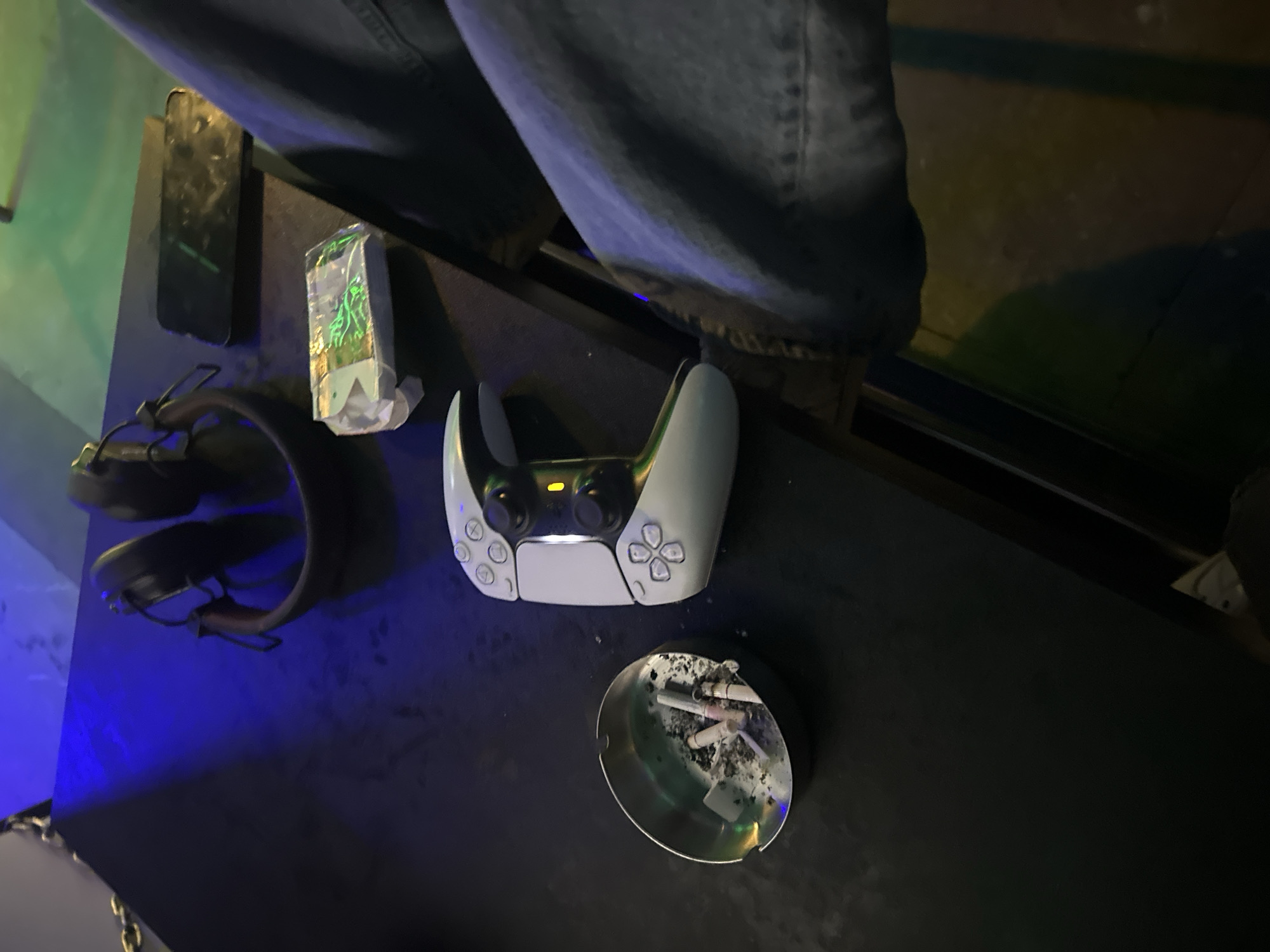 The image shows a black table surface with several items arranged on it. A pair of blue jeans is visible along the top edge of the frame.
On the table, there's a black smartphone, a small package, a pair of black headphones, a white PlayStation 5 controller, and a metal ashtray filled with cigarette butts and ash.
The location is not discernible from the image. It is not possible to determine the image location is Yerevan, Armenia.