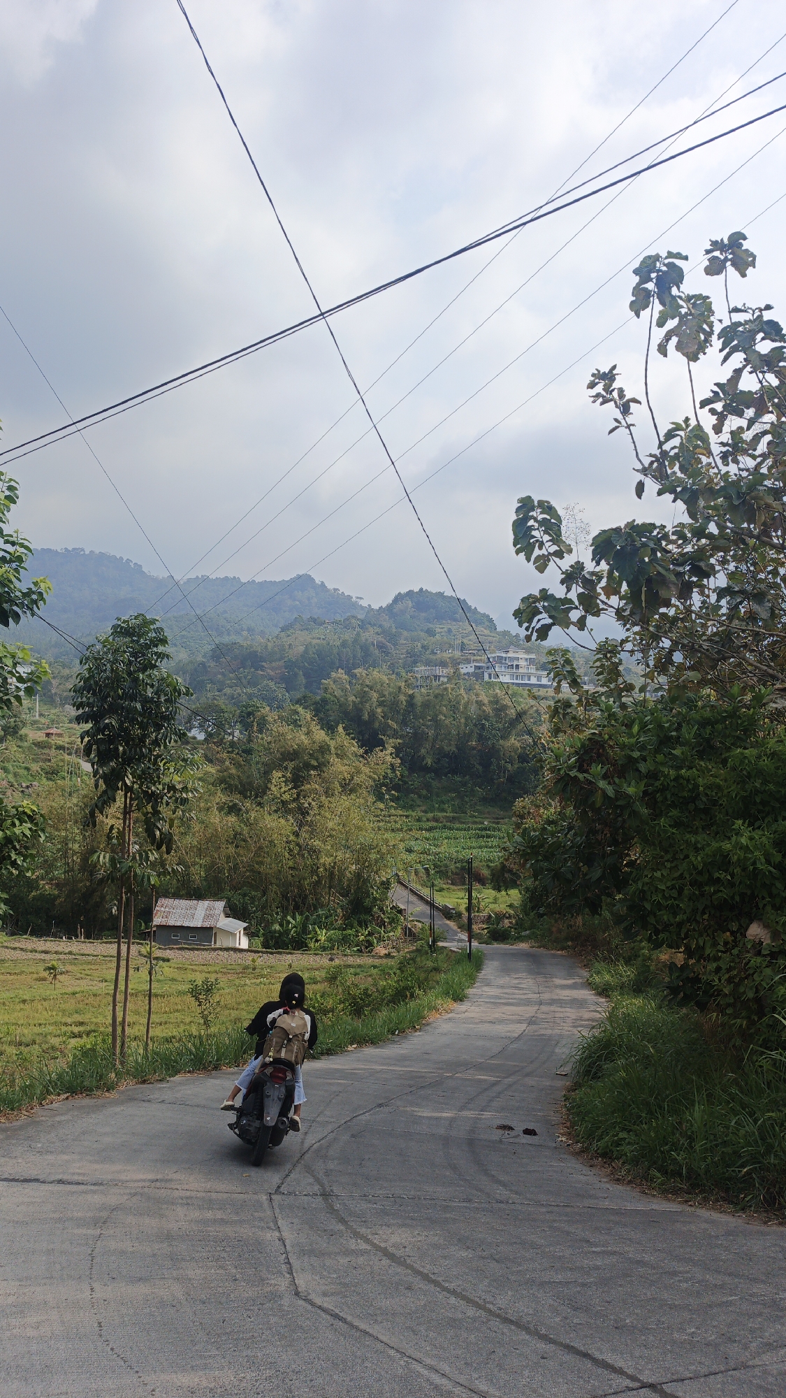 The image depicts a rural scene on a paved road in Sajen, Indonesia. A person is riding a motorcycle with another passenger on the back, both wearing casual clothing and backpacks. The road curves to the right, with lush green vegetation lining both sides. In the background, rolling hills covered in trees and some scattered buildings are visible under a cloudy sky. Power lines crisscross the sky. To the left of the road, there is a small building with a metallic roof and a field of crops. The overall atmosphere is serene and indicative of a natural landscape.
