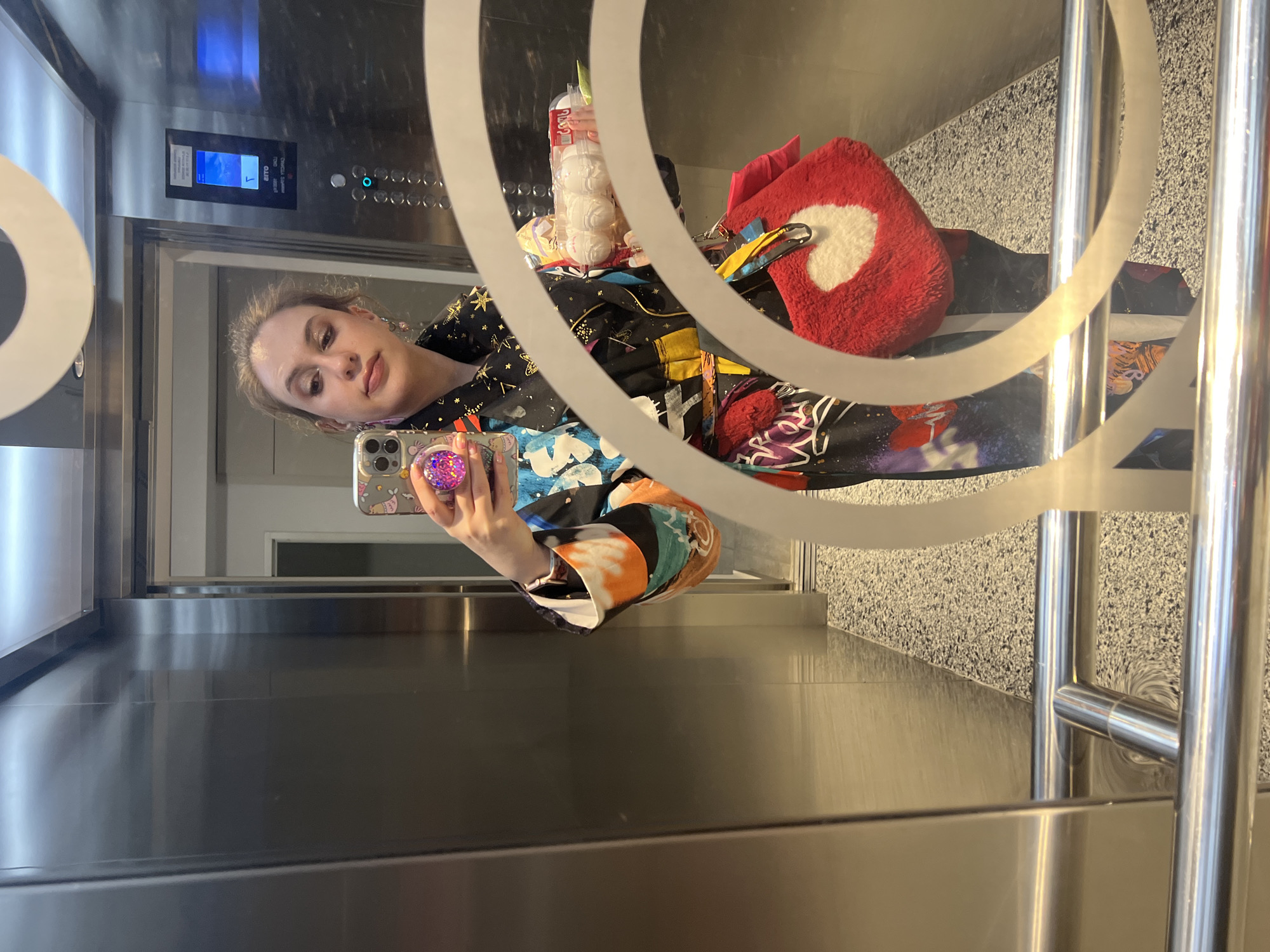 A young woman is captured in a mirror selfie, likely taken inside an elevator. She stands in the reflection, holding a smartphone with a prominent, glittery pink pop socket, appearing to look at her phone's screen as she photographs herself. Her light brown hair is styled back, and she wears makeup. She is dressed in a distinctive black jacket or top, adorned with vibrant, multi-colored patterns including stars and abstract designs.
Beside her in the reflection are several items: a bright red, fluffy object resembling a heart-shaped pillow or bag, and a clear package containing multiple white, spherical items, possibly bath bombs or similar cosmetic products.
The setting is a modern elevator, characterized by sleek metallic walls and a reflective surface that serves as the mirror. An elevator control panel is visible, featuring various buttons and a digital display showing blue and white graphics, along with some mirrored text that appears to read "OTTO". The floor reflected in the mirror has a speckled, granite-like texture. Decorative, curved white lines are present on the reflective surface, adding to the visual composition. The overall impression is a personal moment captured within a contemporary urban environment. This scene takes place in Tbilisi, Georgia.