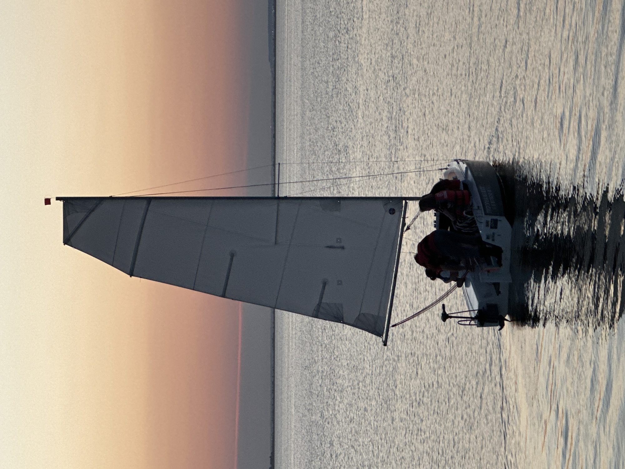 A sailboat with a white sail navigates calm waters during sunset. The sky displays a gradient of soft orange and pink hues. Onboard the sailboat, several individuals are visible, some wearing life vests. The boat's reflection shimmers on the water's surface. The horizon shows a distant landmass. The scene is set on Lake Borovoe, located in the Burabay National Park near Kokshetau, Kazakhstan.