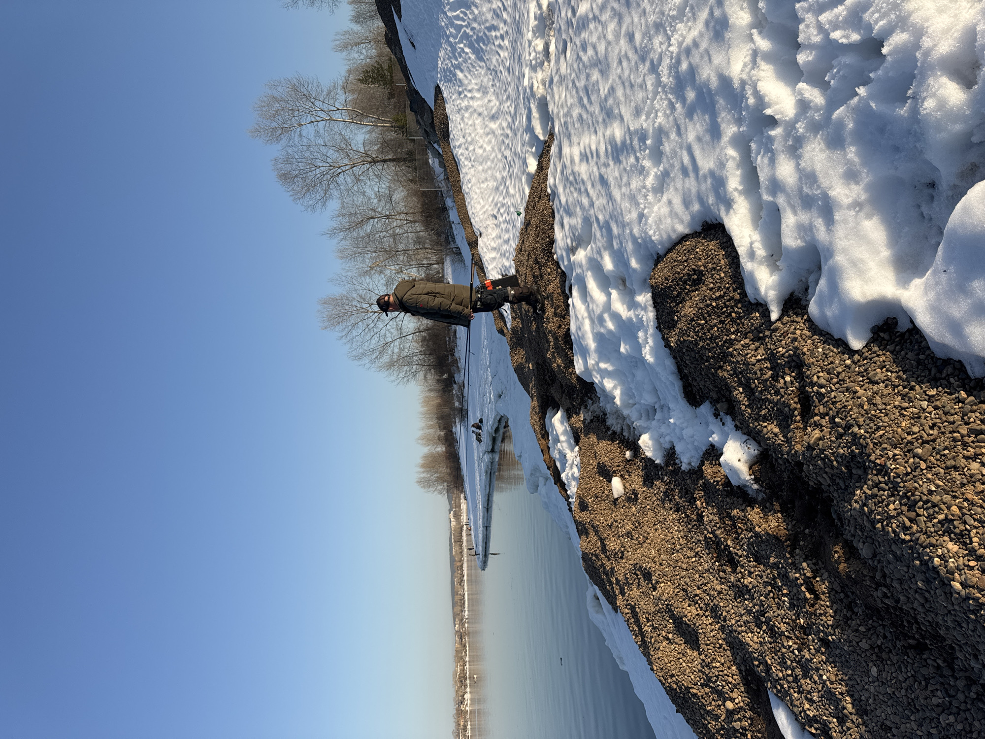 A person stands on a shoreline in Chaikovsky, Russia. The shore is covered in rocks and patches of snow. Behind the person, bare trees line the edge of the shore. The person is wearing a dark coat and hat. In the distance, a body of water reflects the clear blue sky. Buildings are visible on the opposite shore.