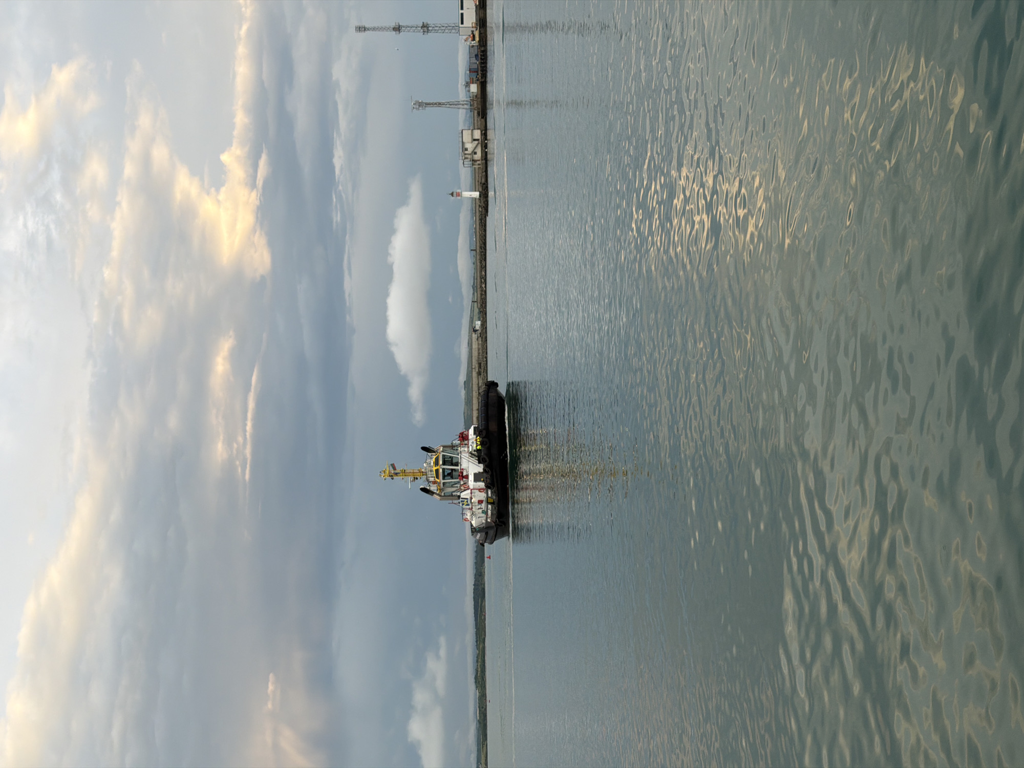 This image captures a tranquil port scene in Burgas, Bulgaria, likely during the golden hour of either sunrise or sunset, given the soft, warm light on the clouds and water. The composition is oriented vertically but depicts a horizontal landscape.
The main subject is a white and yellow tugboat, appearing stationary or docked near a dark pier or breakwater in the mid-ground. The vessel features a prominent yellow superstructure and a white hull.
To the right, a significant portion of the image is filled with calm, rippling water, reflecting the sky's light in shimmering patterns. The water's surface shows intricate textures and movements, with golden hues suggesting the sun's indirect influence.
Along the distant horizon, a narrow strip of land or coastline extends, punctuated by a small white lighthouse with a red cap. Further along the coast, various industrial structures, including tall cranes or communication masts, are visible, signifying an active port area.
The sky above is partly cloudy, with large, diffused clouds showing soft, warm illumination, particularly on the left side. The lighting creates a serene atmosphere, emphasizing the quiet beauty of the working port. No people are visible in the scene, and no discernible text can be read on any of the boats or structures.