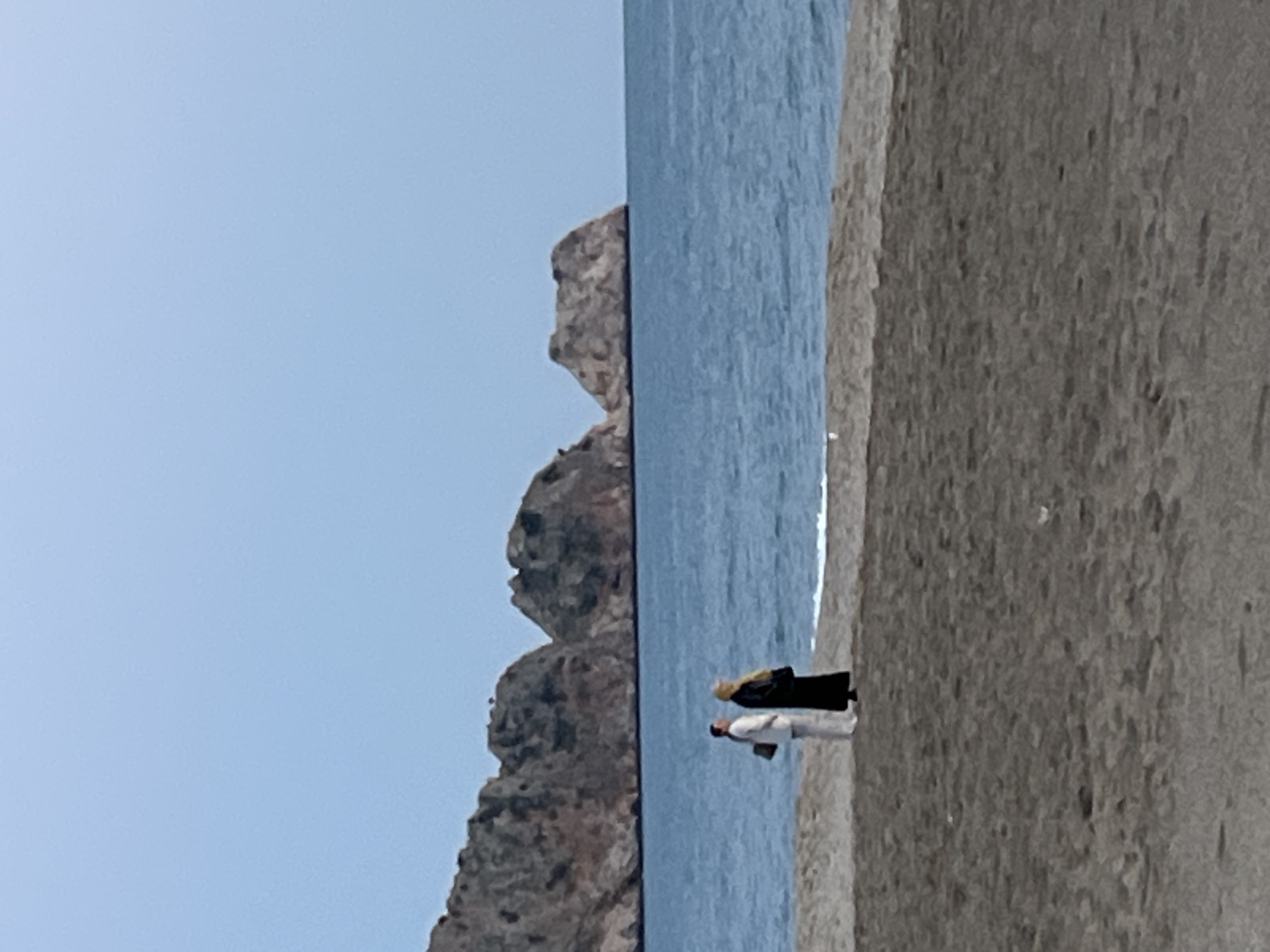 Two people are walking along a beach in Alanya, Turkey. A rock formation is to the left of the frame. The sky is clear. The beach is sandy. The water is calm.