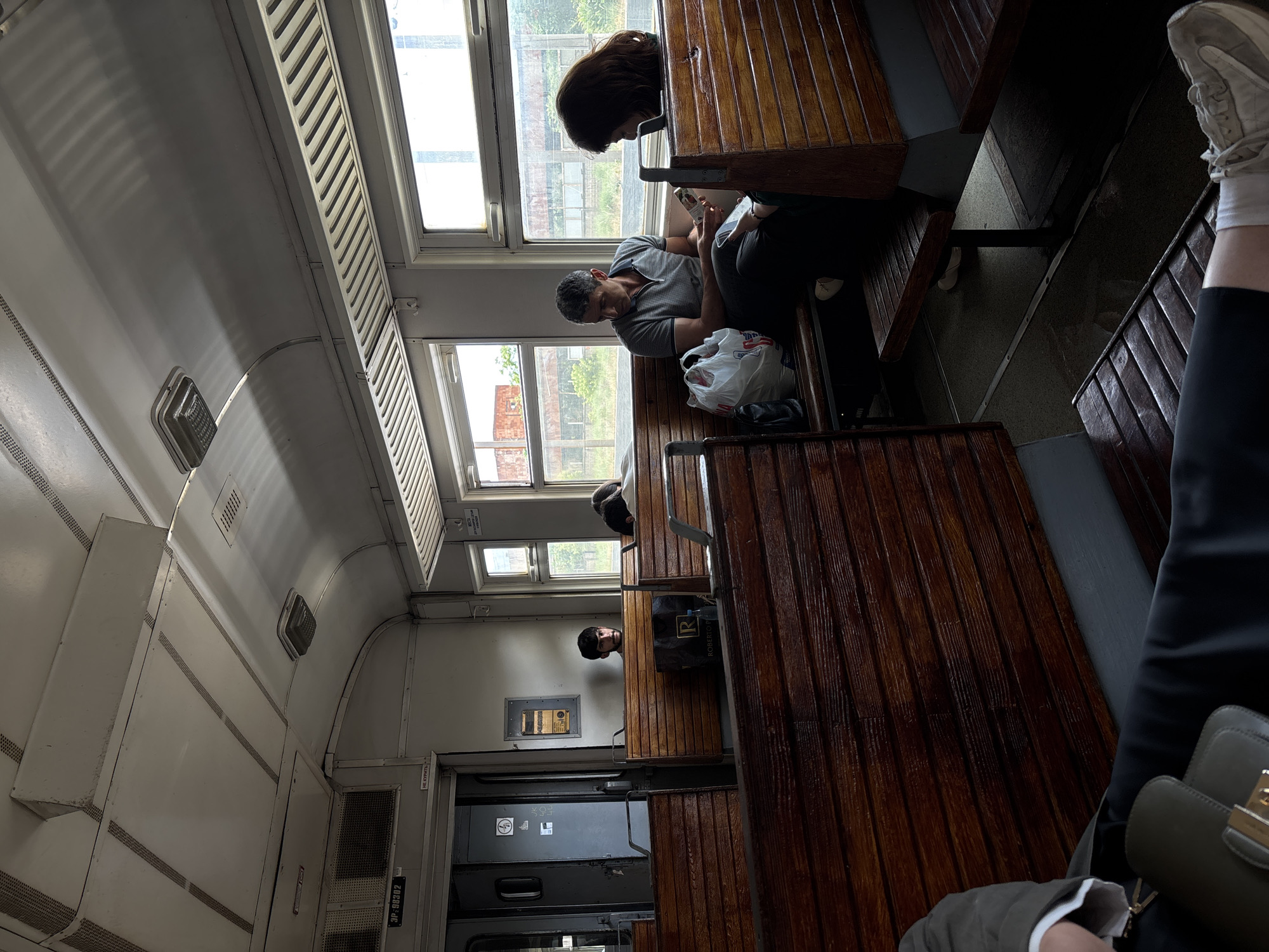 The image depicts the interior of a train carriage, likely on a public transport route, featuring rows of wooden benches. Several passengers are seated. In the foreground, a person's legs and feet, clad in athletic shoes and white socks, are visible, suggesting they are seated or reclining. Further back, a man in a grey polo shirt sits next to a woman with dark hair, both looking at their phones. Between them is a white plastic bag. Another passenger with dark hair is seated further down the carriage. The train carriage appears older, with visible lighting fixtures and metal accents on the ceiling and windows. The windows offer a view of an outdoor environment with greenery and what appears to be industrial or abandoned structures. The overall lighting suggests it is daytime. The provided information suggests the location is Yerevan, Armenia, implying this is a train within or connected to the city.