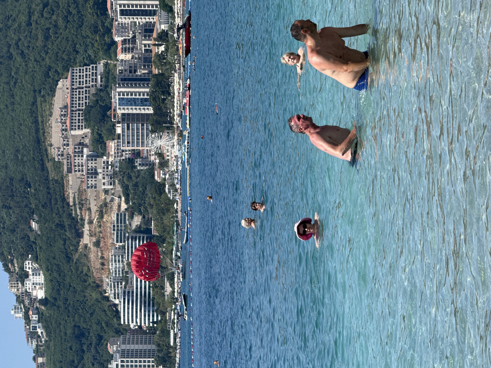 The image depicts a lively scene at a beach in Boreti, Montenegro. The foreground is dominated by the clear, turquoise waters of the Adriatic Sea, where several people are enjoying a swim. Two men are prominently visible in the water, with one facing the camera and the other with his back to it. Further out, a woman with blonde hair is also in the water, and another person wearing a pink hat is visible.
In the background, a bustling shoreline is lined with beachgoers and various watercraft. A vibrant red parasail hangs suspended in the air above the water, hinting at recreational activities. Beyond the beach, a dense green hillside rises steeply, dotted with numerous multi-story buildings, suggesting a developed coastal town. The architecture appears modern, with many buildings featuring uniform window patterns. A Ferris wheel is also visible in the distance, adding to the sense of a tourist destination. The overall atmosphere is bright and sunny, indicative of a pleasant summer day. The image captures a moment of leisure and recreation in a scenic coastal setting.
