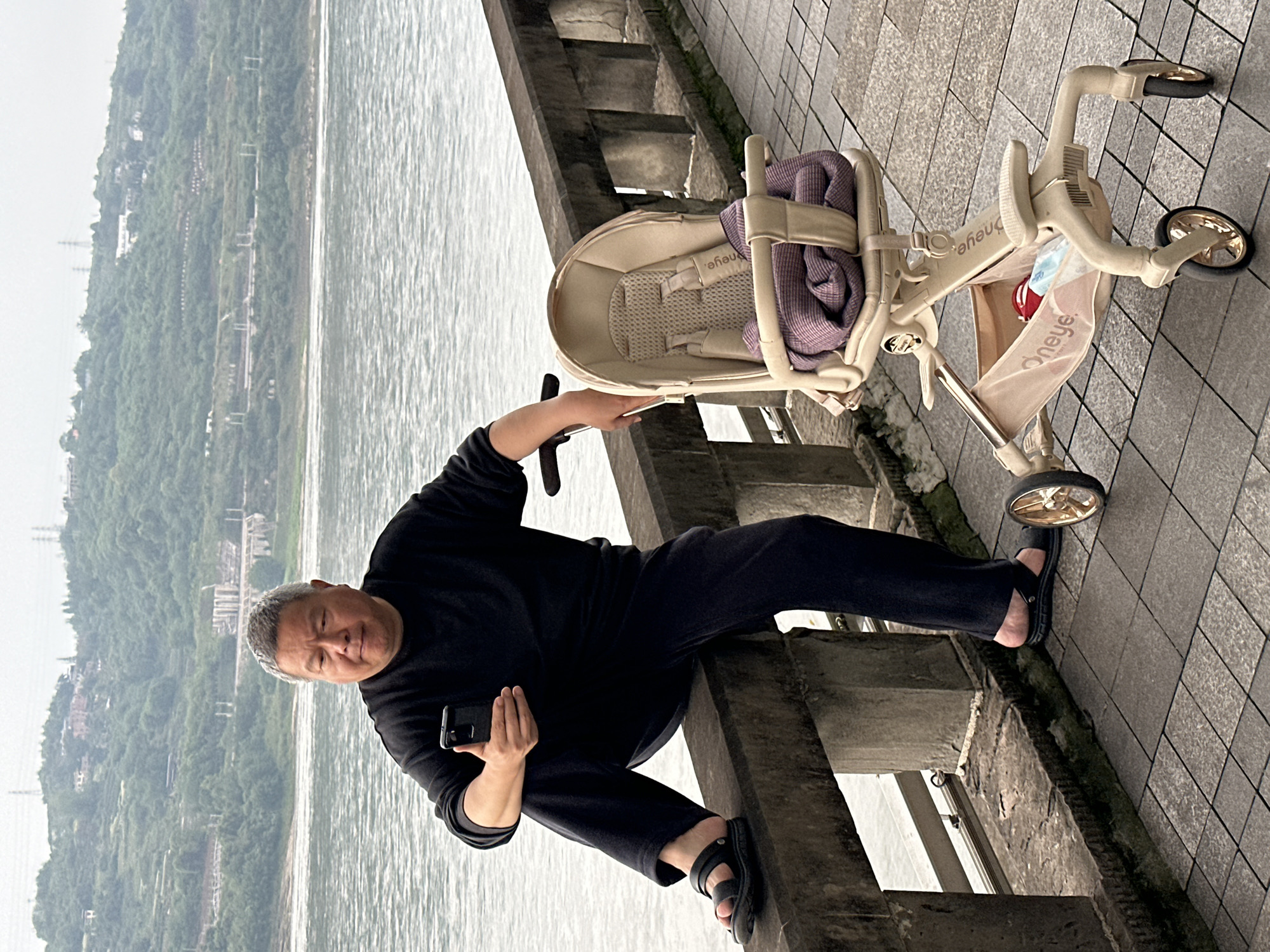 A man with grey hair is sitting on a grey concrete ledge near a body of water in Jiangjin, China. He is wearing black clothing and black sandals. He is holding a smartphone in his right hand and looking at the camera. To the right of the man is a beige stroller or tricycle with a purple blanket inside. The background shows a body of water and a green hillside. The environment appears to be a grey, overcast day.