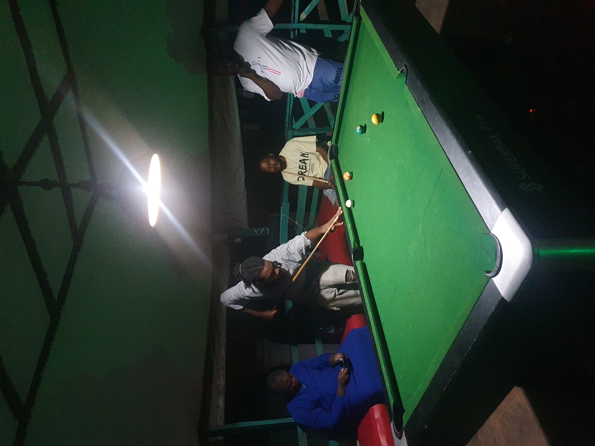The image depicts an indoor scene where a group of four men are gathered around a pool table. The pool table is green with several balls on it, including a yellow ball, a blue ball, an orange ball, and a white cue ball. One man, wearing a striped shirt and ripped jeans, is actively playing pool, holding a cue stick and aiming at the white ball. He has dreadlocks and is facing the pool table. To his left, a man in a blue robe is seated and looking at his phone. To the right of the playing man, another man is standing and observing the game, wearing a beige t-shirt with the word "DREAM" printed on it. Further to the right, a fourth man is standing, wearing a white t-shirt with text that appears to be "PARIS" and blue shorts. The environment appears to be a dimly lit entertainment space with a green ceiling and exposed beams. The lighting is provided by a bright overhead lamp. The pool table has "SUPERMAX PRO" written on its side. The scene is consistent with a pool hall or a recreational area in Lokoja, Nigeria, where individuals are engaged in leisure activities.
