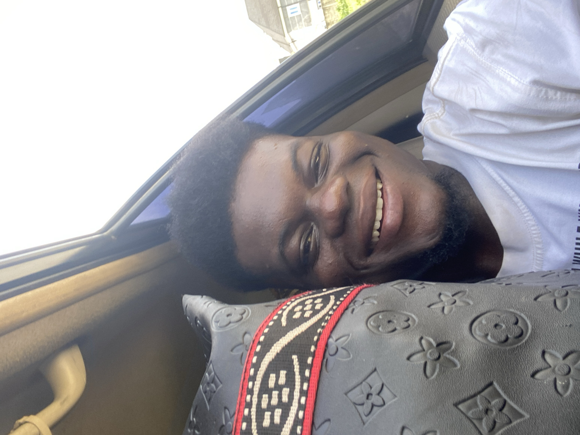 A close-up, portrait-oriented image, rotated 90 degrees clockwise, captures a young, dark-skinned man smiling broadly and looking directly at the viewer. He has short, curly dark hair and a light beard. The man is wearing a white short-sleeved t-shirt.
He is resting his head on a black pillow or cushion adorned with embossed floral and quatrefoil patterns, reminiscent of the Louis Vuitton monogram. A decorative strap, featuring black, red, and off-white geometric patterns, runs diagonally across the pillow.
The man appears to be inside a vehicle, with a window and its frame visible behind him. The bright, almost white light streaming through the window suggests it is daytime. Outside, a blurred view of a building with a dark roof and some green foliage can be seen against the bright sky. The interior of the vehicle, including what appears to be a light brown or beige panel, is visible around the man and the pillow. The man's expression conveys happiness and relaxation. No discernible text is visible in the image.