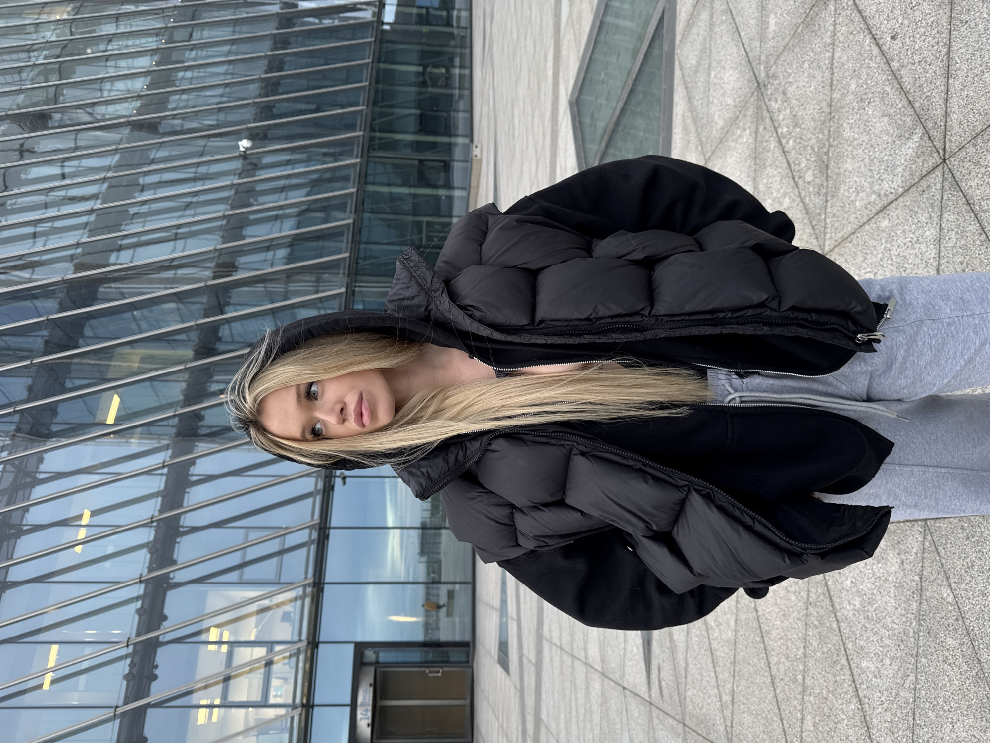 A young woman with long blonde hair and light blue eyes is posing for a photo in an urban setting. She is wearing a black puffer jacket over a black hoodie and light grey sweatpants. Her hair is partially covered by the hood of her jacket. She is looking directly at the camera with a neutral expression.
The background features a modern glass building with a reflective facade, indicating an urban environment. The ground is paved with grey tiles laid in a geometric pattern. The lighting suggests it is daytime, with soft, diffused light. The overall mood is casual and relaxed.
The image is oriented vertically. The location is Saint Petersburg, Russia. No visible text is present in the image.