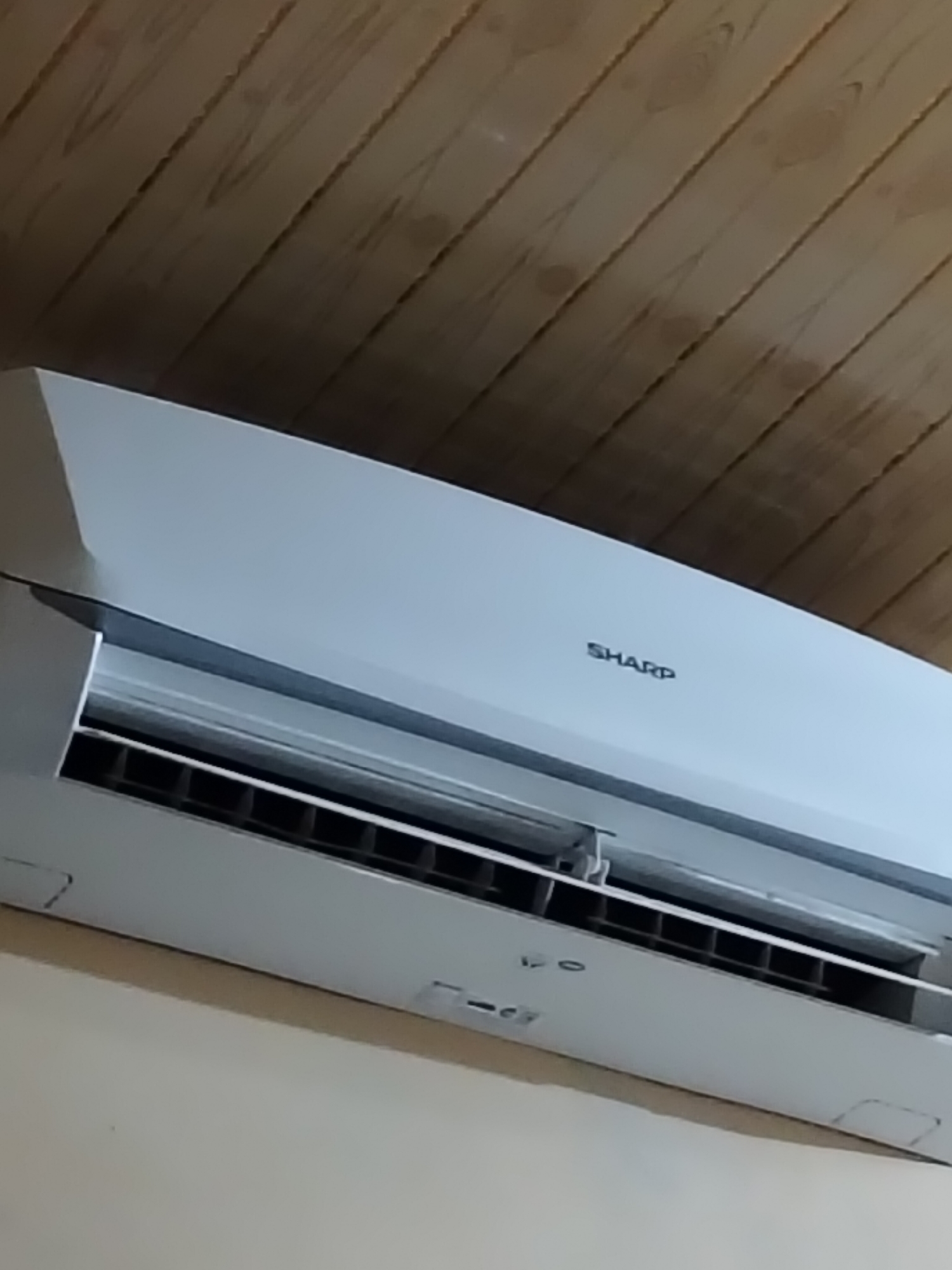 The image shows a close-up of a white Sharp air conditioning unit mounted on a light-colored wall. The unit has a vent with horizontal slats. Above the air conditioner, there is a wooden ceiling with diagonal planks. The image is taken from a low angle, looking upwards. There is no indication of people or any specific location within the city of Payagaboh, Indonesia.