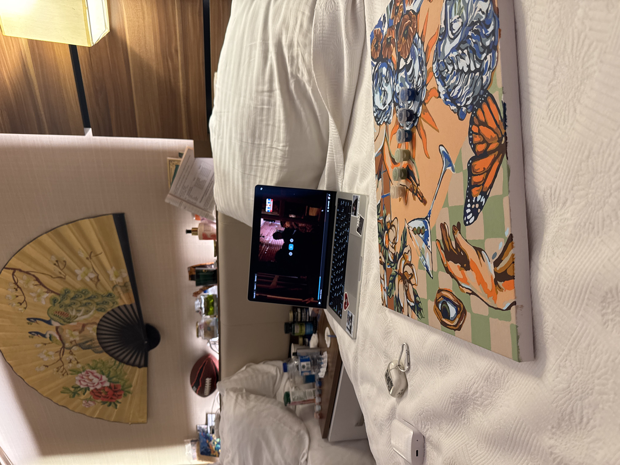 The image shows a bed with a laptop open and a painting kit on it. The laptop displays a video. Nearby, there are various items on a nightstand, including a football, jars, and bottles. A decorative fan is mounted on the wall behind the bed. The scene appears to be in a hotel room. The location is not specified in the image or provided context.