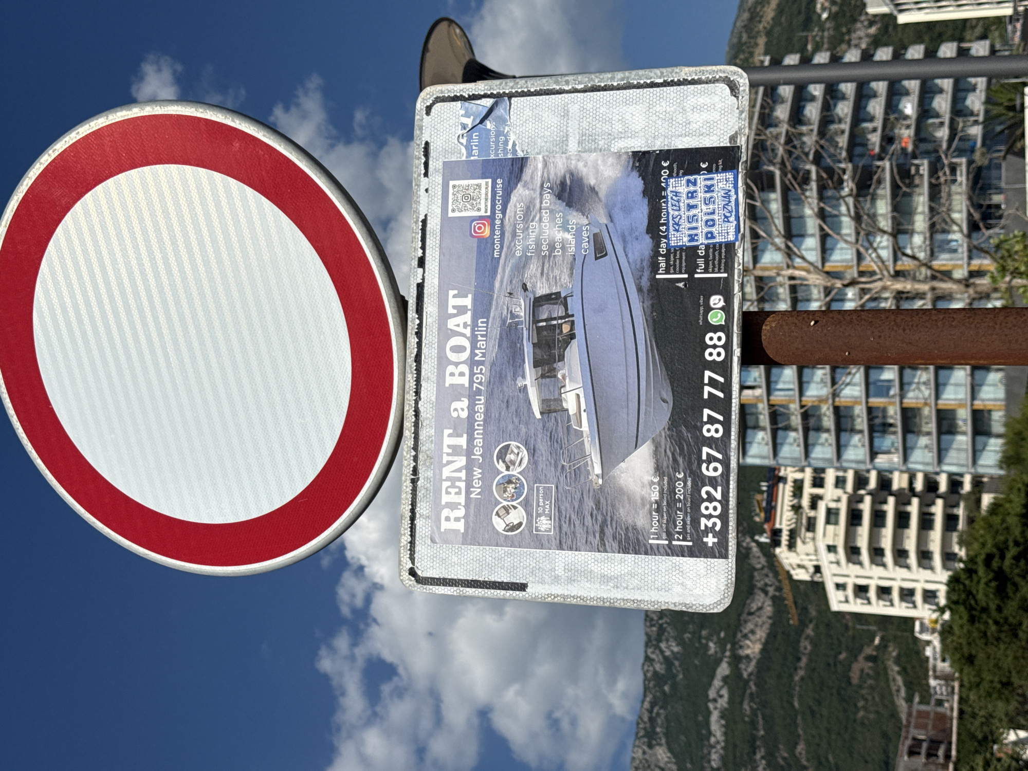 This media file is a photograph taken outdoors, likely during the daytime given the bright sky and visible clouds. The scene features a red and white circular road sign in the foreground on the left, which is partially obscured. Attached to a metal pole behind the sign is a flyer for a boat rental service. The flyer prominently displays the text "RENT a BOAT" and mentions "New Jeanneau 795 Marlin." It also lists various activities such as "excursions," "fishing," and "secluded bays," along with pricing for 1-hour and 2-hour rentals.
Further details on the flyer include an Instagram handle "@MONTENEGROCRUISE" and a phone number "+382 67 87 77 88." There are small icons suggesting features like a 10-person maximum capacity and some sort of Bluetooth connectivity or speaker. A partial view of a modern apartment building or hotel complex is visible in the background, set against a backdrop of a steep, forested mountainside. The overall impression is an advertisement for leisure activities in a scenic coastal area. There are no people clearly depicted in the image. The weather appears to be clear with some scattered clouds. The presence of the road sign suggests a public space, possibly near a waterfront or a road leading to one.