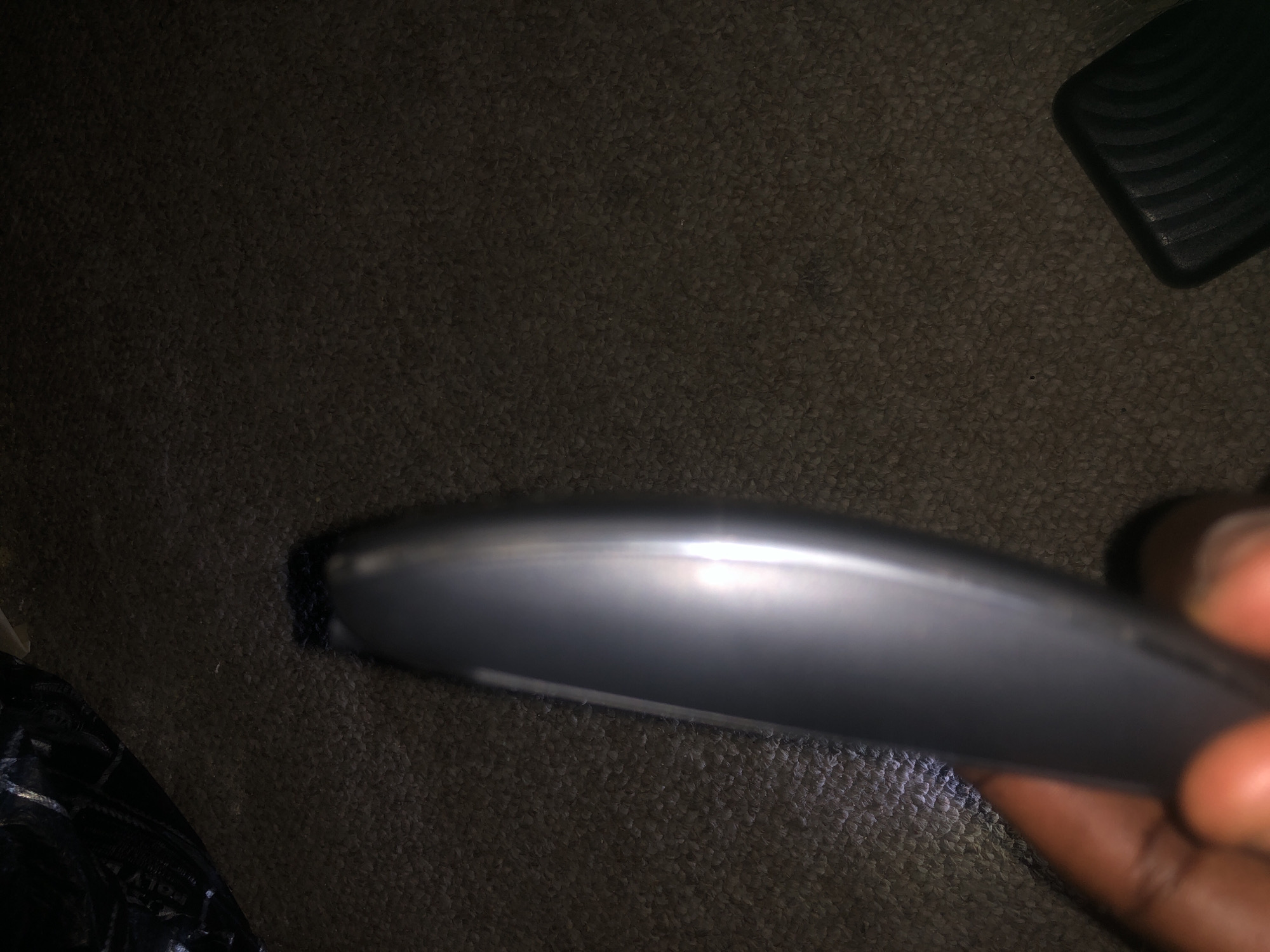 A hand is holding a silver object that appears to be a remote control or a similar electronic device. The object is oriented vertically in the frame, with the hand gripping its lower half. The background consists of a dark, textured surface, possibly carpet or upholstery, with a black object with a ridged pattern visible in the lower right corner. The lighting in the image is dim, with a strong reflection on the silver object. The scene is not clearly indicative of any specific location within Jalingo, Nigeria, or any other city.