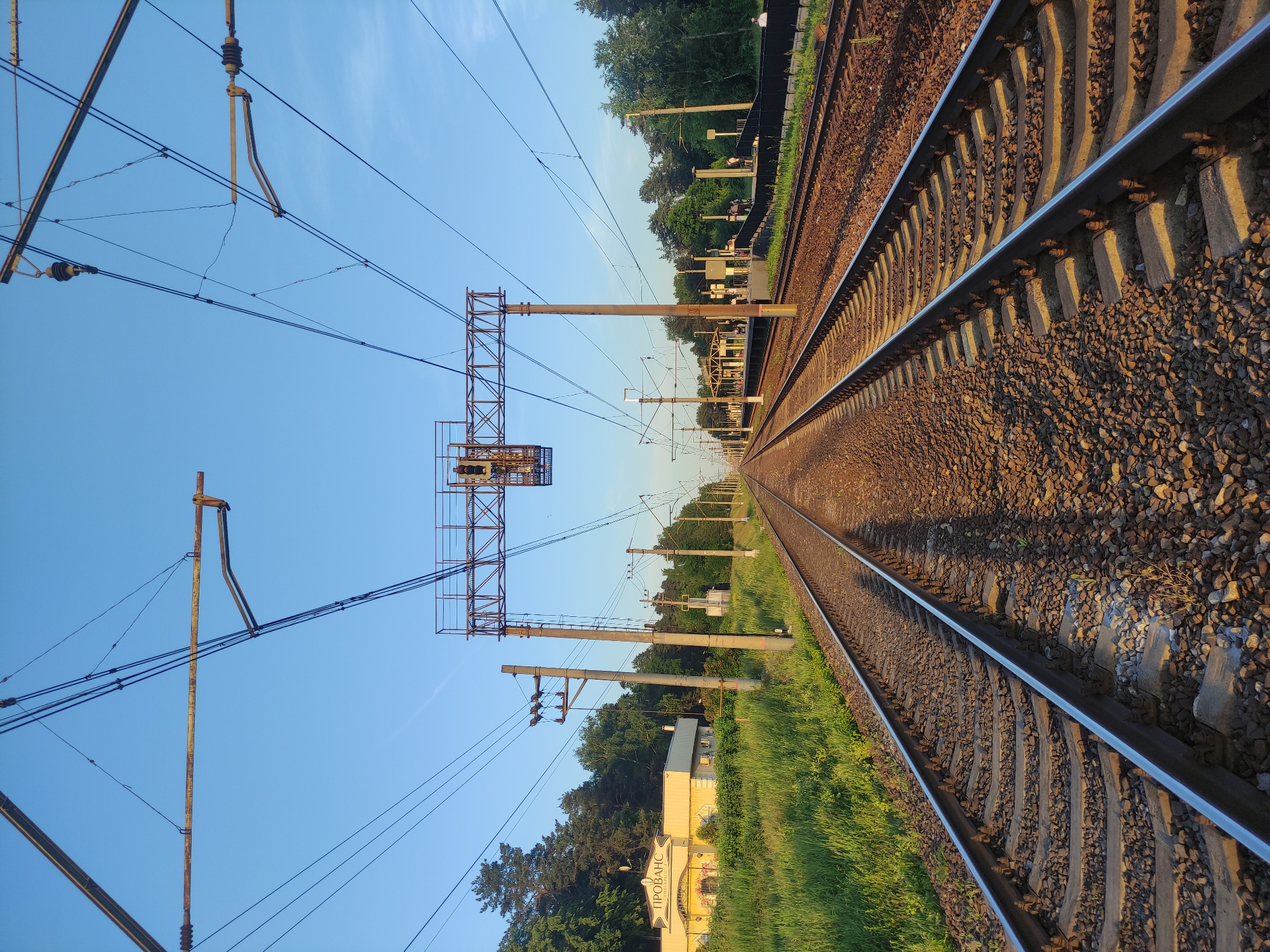 The image depicts a multi-track railway line in Kratovo, Russia, observed from a ground-level perspective during golden hour lighting, casting long shadows. Two primary tracks, constructed with metal rails on gravel ballast and sleepers, extend linearly into the distant horizon.
An elaborate overhead catenary system spans the tracks, featuring concrete support poles, horizontal metal gantry structures, and numerous electrical wires. A prominent railway signal gantry, located centrally above the tracks, displays at least two signal heads, with visible red and yellow aspects on the left-most signal.
On the left side of the tracks, a light-yellow building with a dark roof is visible, bearing the Cyrillic inscription "ПРОВАНС" (Provans/Provence). This building is set against a backdrop of dense green trees and a field of tall green grass.
On the right side of the tracks, a railway station platform is present, featuring a covered waiting area with supporting pillars. Approximately 5-7 indistinct human figures are standing on the platform. A dark metal fence separates the platform from the adjacent track. Beyond the platform, a thick forest line extends into the background. The sky above is clear and blue with no significant cloud cover. No specific actions or interactions are discernible among the individuals on the platform due to the distance.