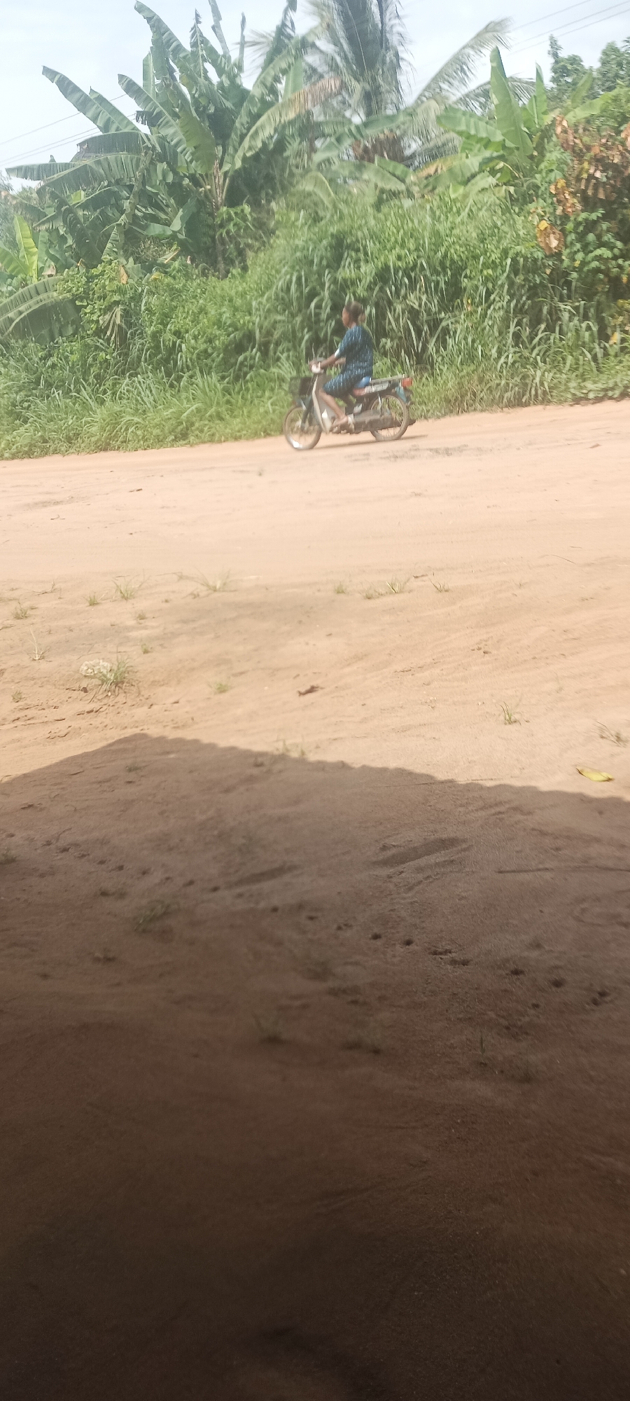 A dirt road scene in Amauzu, Nigeria. A person rides a moped on a dirt road that is bordered by dense green vegetation. The foreground is obscured by a dark shadowed area.