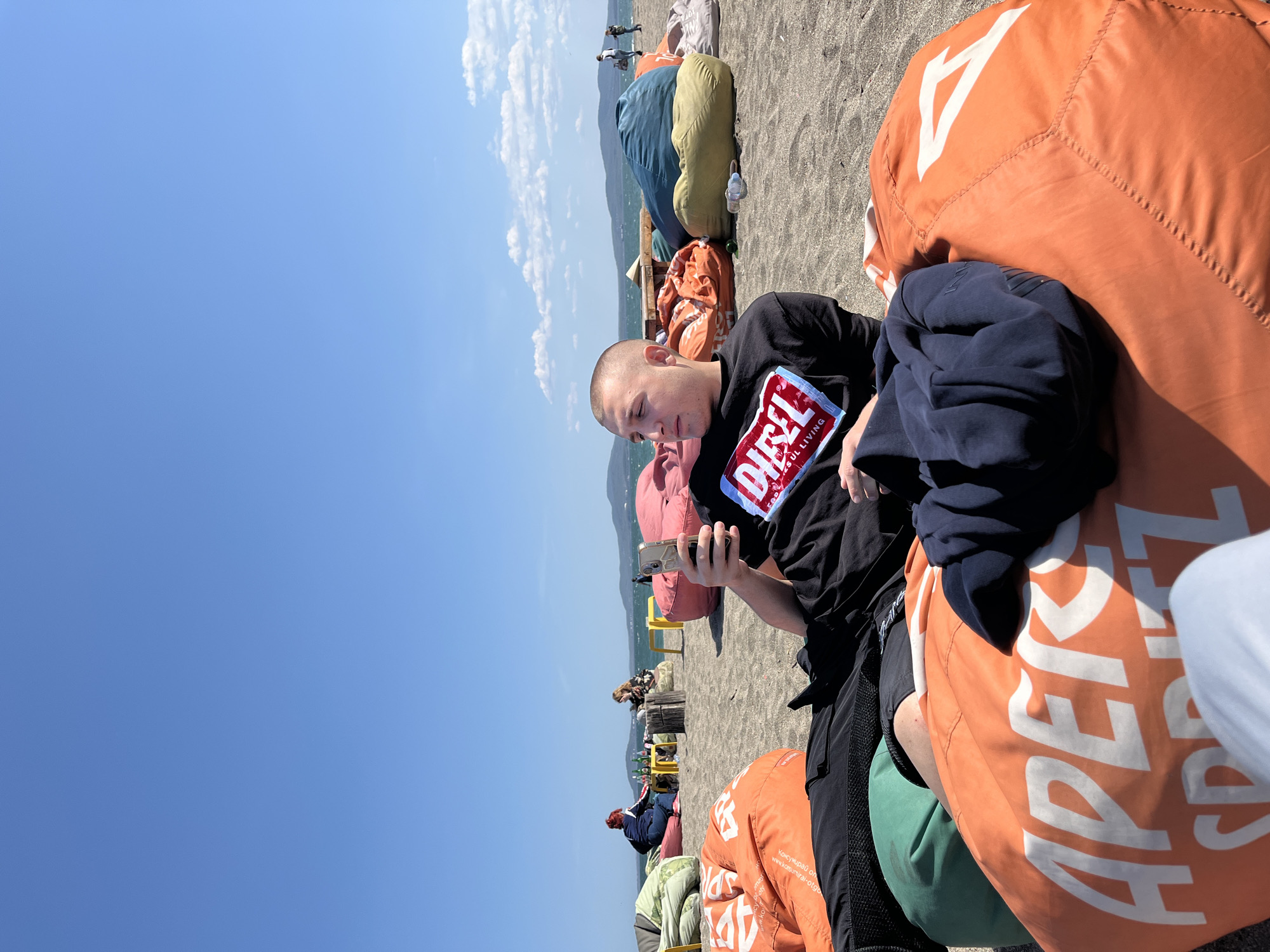 This media file depicts a sunny day on a beach in Burgas, Bulgaria, focusing on a young person relaxing and using a smartphone. The main subject, a male with a shaved head, is seen from a slightly elevated side angle, wearing a black t-shirt featuring a prominent "DIESEL FOR SUCCESSFUL LIVING" logo in red and blue. He is seated comfortably on a large, orange bean bag chair on dark sand, engrossed in his phone. A dark blue sweatshirt or jacket is draped beside him on another part of the bean bag.
The beach is dotted with numerous colorful bean bag chairs in shades of orange, green, pink, blue, and yellow, with other people in the background also relaxing or walking. The orange bean bag chair in the immediate foreground displays large white text, partially visible as "APEROL," and some Cyrillic text below it, "Копируващи от www.kopiriai-otg...".
The setting is an open beach with dark-colored sand, stretching towards the sea which is visible in the middle ground. Distant mountains or hills line the horizon under a clear, bright blue sky with a few scattered white clouds, indicating pleasant weather during the daytime. The overall scene conveys a relaxed, leisurely atmosphere on a vibrant beach.