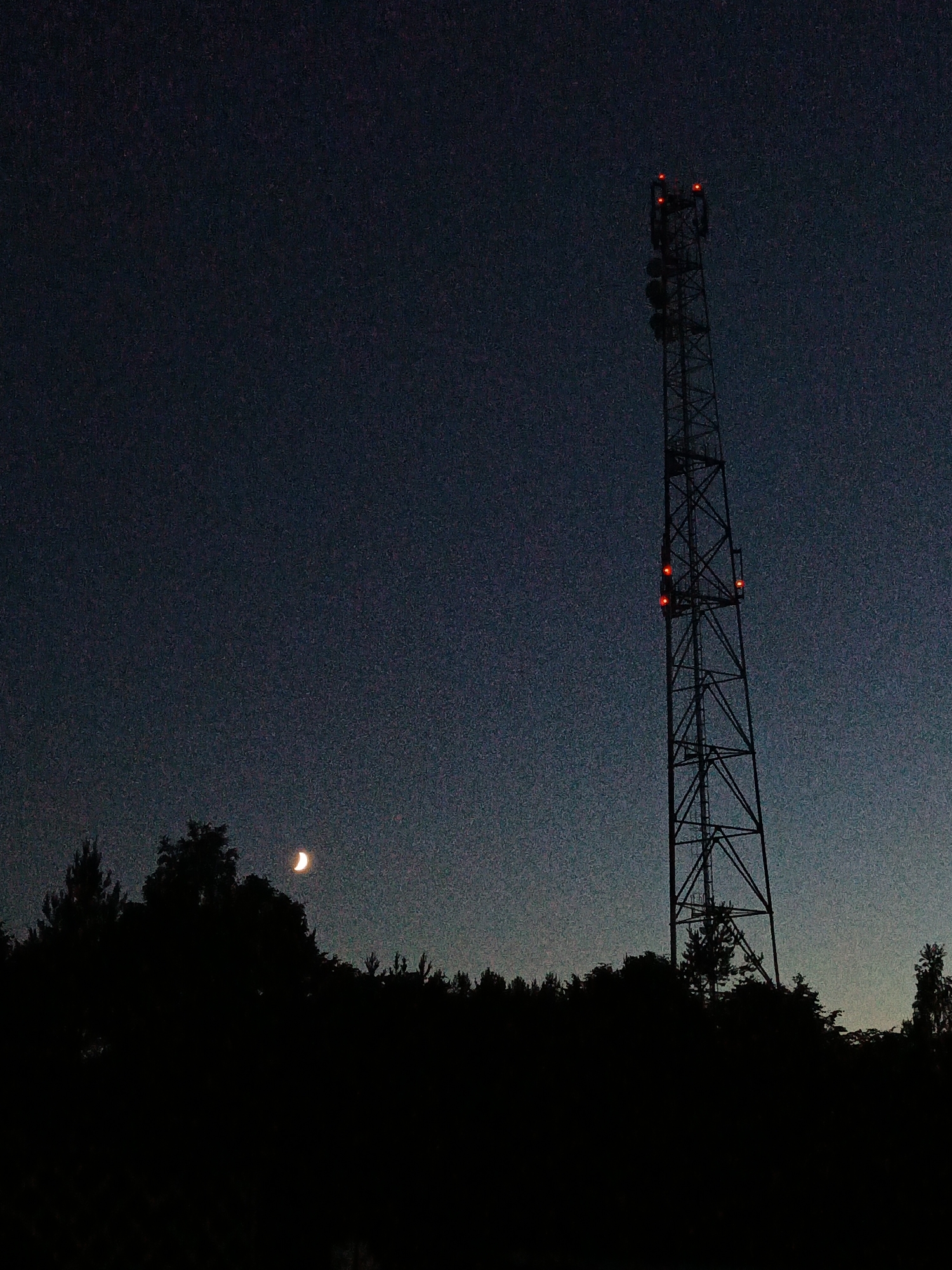 This is a low-light, full-frame photograph taken at dusk in the city of Porechye, Belarus. The image features a tall, dark telecommunications tower with red lights illuminating its upper section, standing in silhouette against a deep blue, starry sky. In the lower left portion of the frame, a sliver of the crescent moon is visible above a dark, silhouetted treeline that stretches across the bottom of the image. The landscape appears to be rural or semi-rural, with the dense forest contrasting against the man-made structure of the tower. No people are visible in the scene.