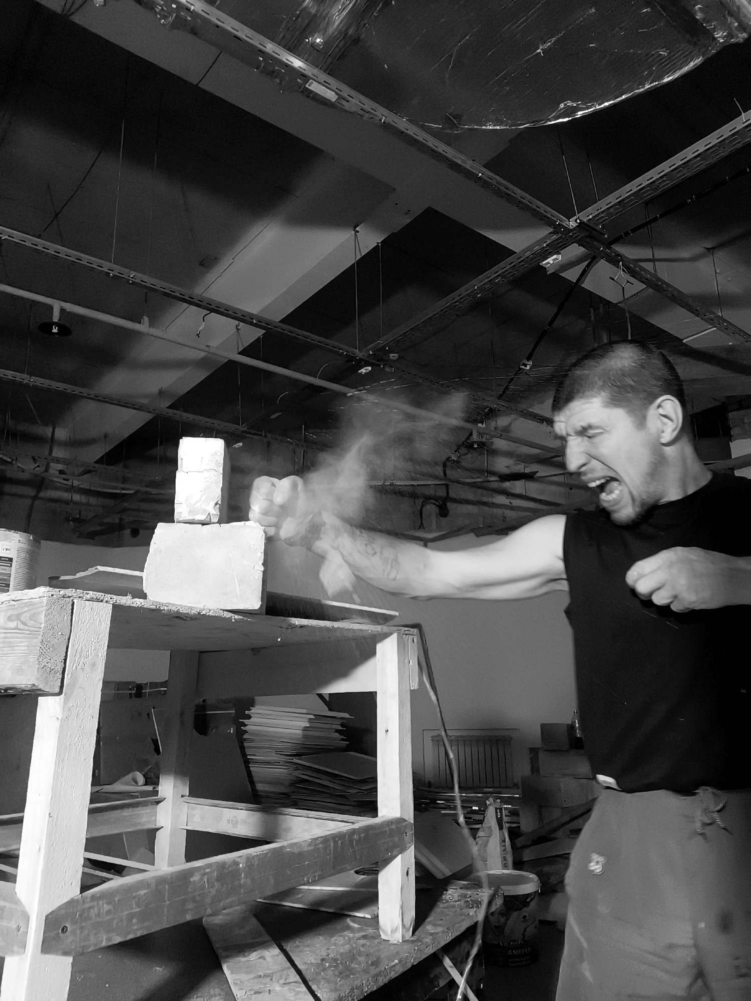 In a black and white photograph, a man with short hair and a muscular build is captured in mid-action, punching a stack of white objects, possibly bricks, that are balanced on a wooden surface. His face is contorted in an expression of exertion, with his mouth open in a shout, and dust or debris is flying from the point of impact. He is wearing a dark sleeveless shirt and dark pants. The background appears to be an indoor space with exposed ceilings, pipes, and electrical conduits, suggesting a workshop or unfinished building. The lighting is dramatic, casting strong shadows and highlighting the dust cloud. The scene is devoid of any discernible signage or landmarks that would specifically place it within the city of Qarağandı city administration, Kazakhstan.