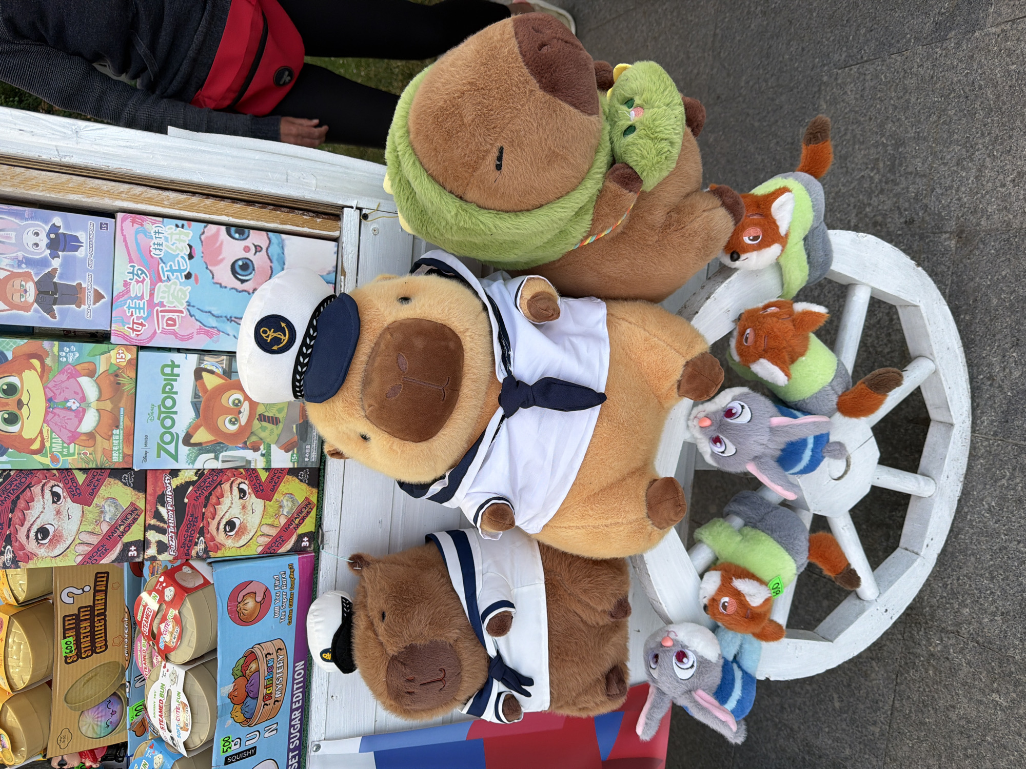 The image displays a street vendor's stall, likely located in Yalta, Ukraine, featuring a collection of plush toys arranged on a white wooden cart. The cart's wheels and frame are visible, and it appears to be positioned on a paved surface, possibly a sidewalk or street. Behind the cart, stacked boxes and merchandise are visible, including what seem to be toys or collectible items.
The main subjects are several large plush capybara toys dressed in sailor outfits. Two are positioned prominently on the cart, one wearing a white sailor shirt with a dark blue tie and cap, and another similarly attired. A third capybara, also a plush toy, is to the right, wearing a green hood and a small green pouch. Smaller plush toys, resembling rabbits and foxes, are arranged on the cart's wheels and base.
In the background, the upper portion of a person is visible, wearing a dark long-sleeved shirt and dark pants, with a red fanny pack worn across their chest. The person's hands are resting on the cart. The overall scene suggests a commercial setting outdoors. The merchandise on the shelves behind the capybaras includes boxes with colorful graphics, some featuring cartoon characters and text in Asian languages, along with labels like "STEAMED BUN" and "ZOOTOPIA." Some items appear to be novelty blind boxes.