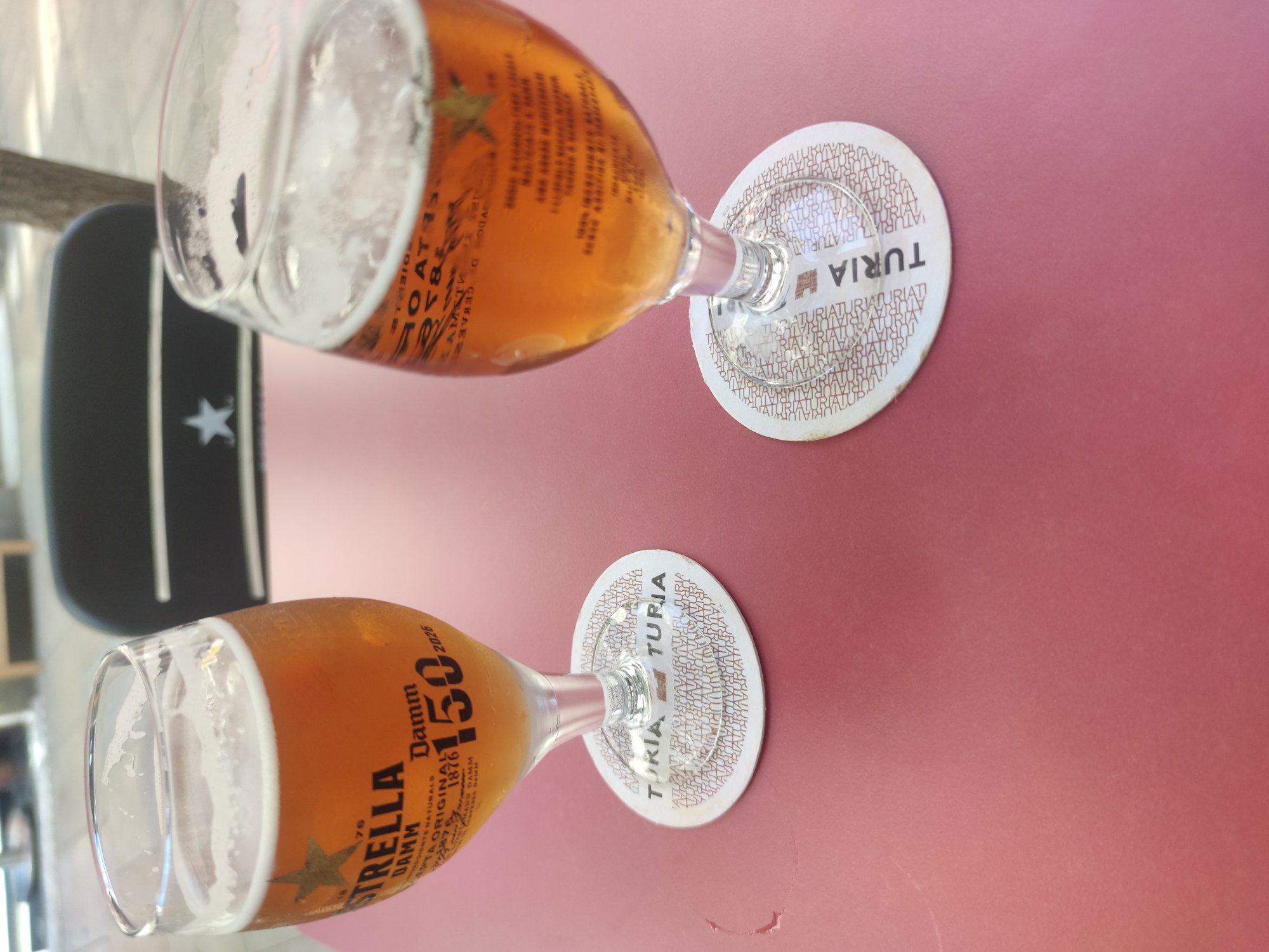 Two glasses of beer sit on a pink table. The glass on the left has a label that reads "Estrella Damm 150 1876 2026". The glass on the right has a label that is partially obscured but appears to be similar. Both glasses are on coasters that read "TURIA". In the background, a dark chair with a white star is visible, along with a tree trunk and a blurred outdoor setting.