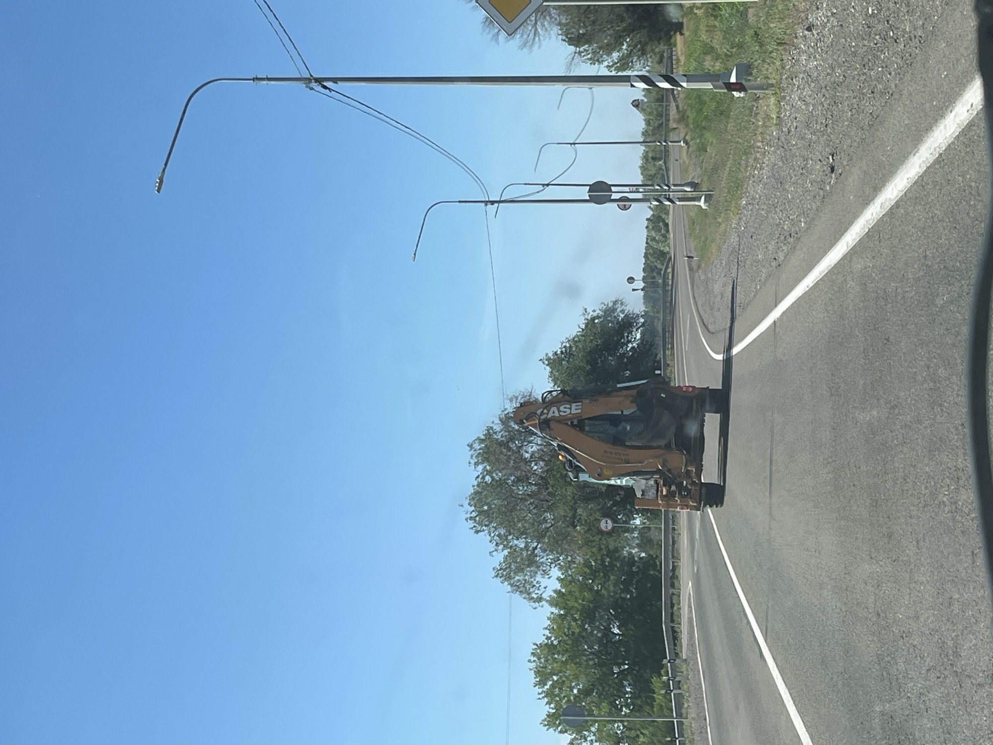 The image depicts a road in Bakhtemir, Russia. A backhoe loader with the brand "CASE" visible on its side is driving on the right side of the asphalt road. The loader is yellow and orange and appears to be a construction vehicle. The road is lined with a gravel shoulder and green trees. In the background, there are utility poles with wires stretching across the sky. The sky is a clear, bright blue, indicating it is daytime with good weather. There are no people visible in the image. The scene suggests a rural or semi-rural setting along a highway.