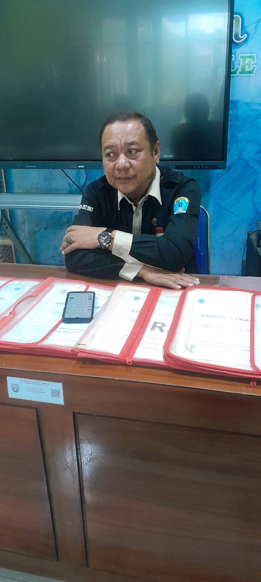 A middle-aged Indonesian man with short, dark hair and a light complexion is seated at a wooden desk. He is wearing a dark long-sleeved shirt with a light-colored collar and a circular emblem on the left sleeve. He has a watch on his left wrist and a ring on his left index finger. His left arm is crossed over his chest, and his right arm is resting on the desk. The man is looking to his left, with his mouth slightly open.
In front of him on the desk are several clear plastic document holders with red trim, each containing papers. A smartphone is placed on top of one of the document holders, displaying a screen with text and icons. Behind the man, a large screen television is visible, displaying a fuzzy image. The wall behind the television has a blue, textured pattern.
A small white sticker with text and a QR code is affixed to the front of the desk below the document holders. The setting appears to be an indoor office or meeting room, possibly in the city of Tahunan, Indonesia, given the context provided by the sticker which reads "PEMERINTAH KABUPATEN JEPARA" (Jepara Regency Government), as Tahunan is a district within Jepara Regency.