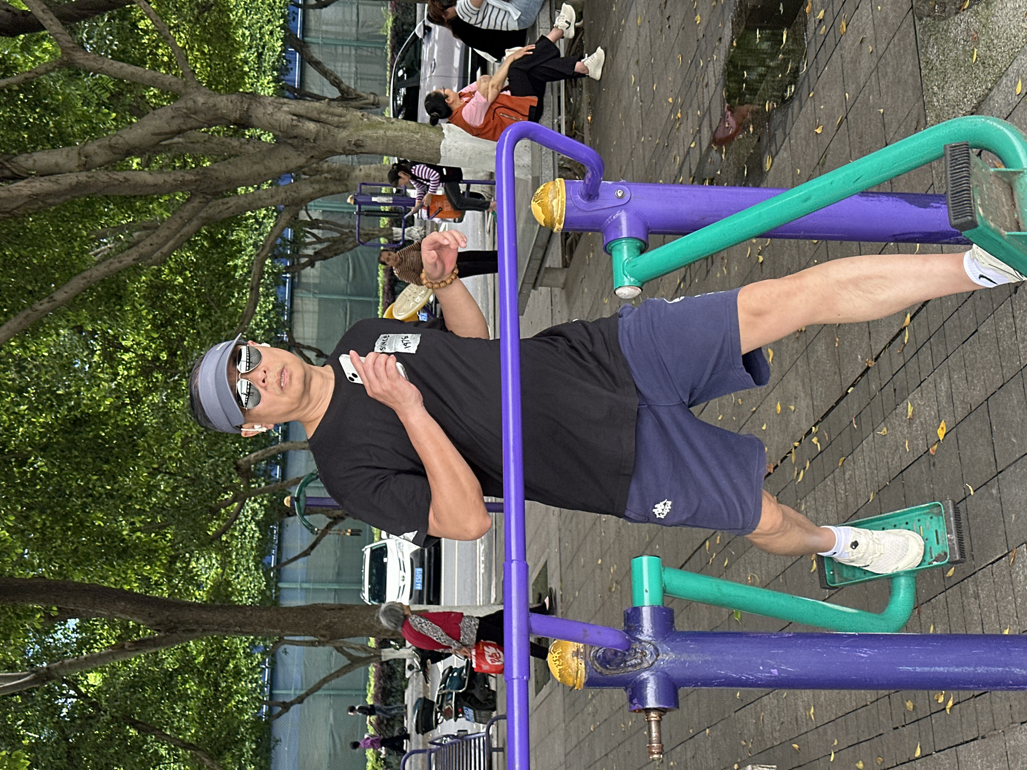 A man exercises on a leg press machine in Jiangjin, China. He wears a visor, sunglasses, a black t-shirt, and blue shorts, and uses a phone. Other people and vehicles are in the background, with trees overhead. The leg press machine is purple and green. The ground is paved.